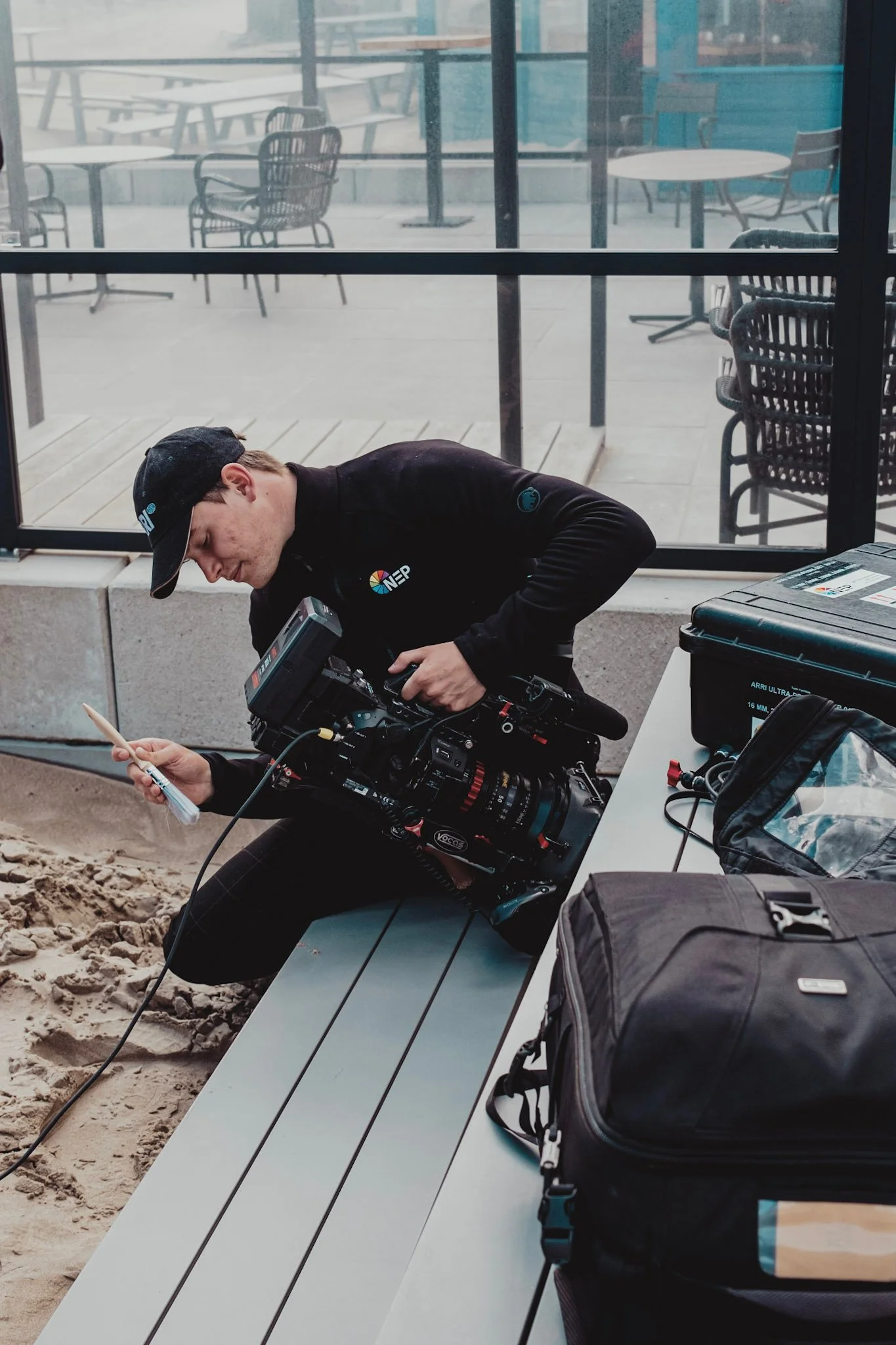 A technician working with professional camera equipment on a gravel surface near an outdoor seating area with glass walls and empty tables and chairs.