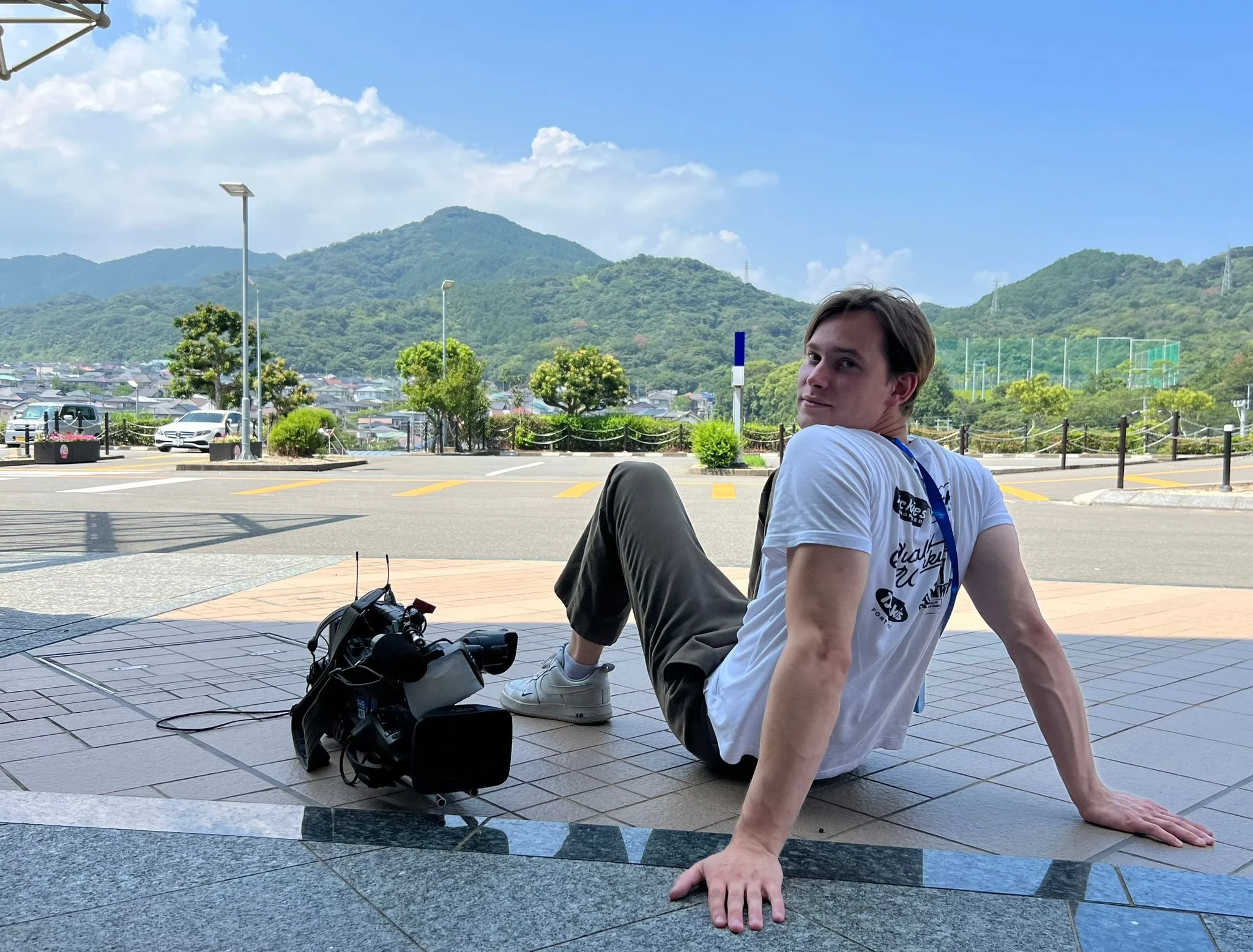 A young man with short brown hair and light skin is sitting on the ground, leaning back on his hands, with his legs bent. He is wearing a white t-shirt and dark pants, and is outdoors with a scenic backdrop of green mountains, trees, and a partly cloudy blue sky. There are street lamps, cars, and a parking area visible behind him.