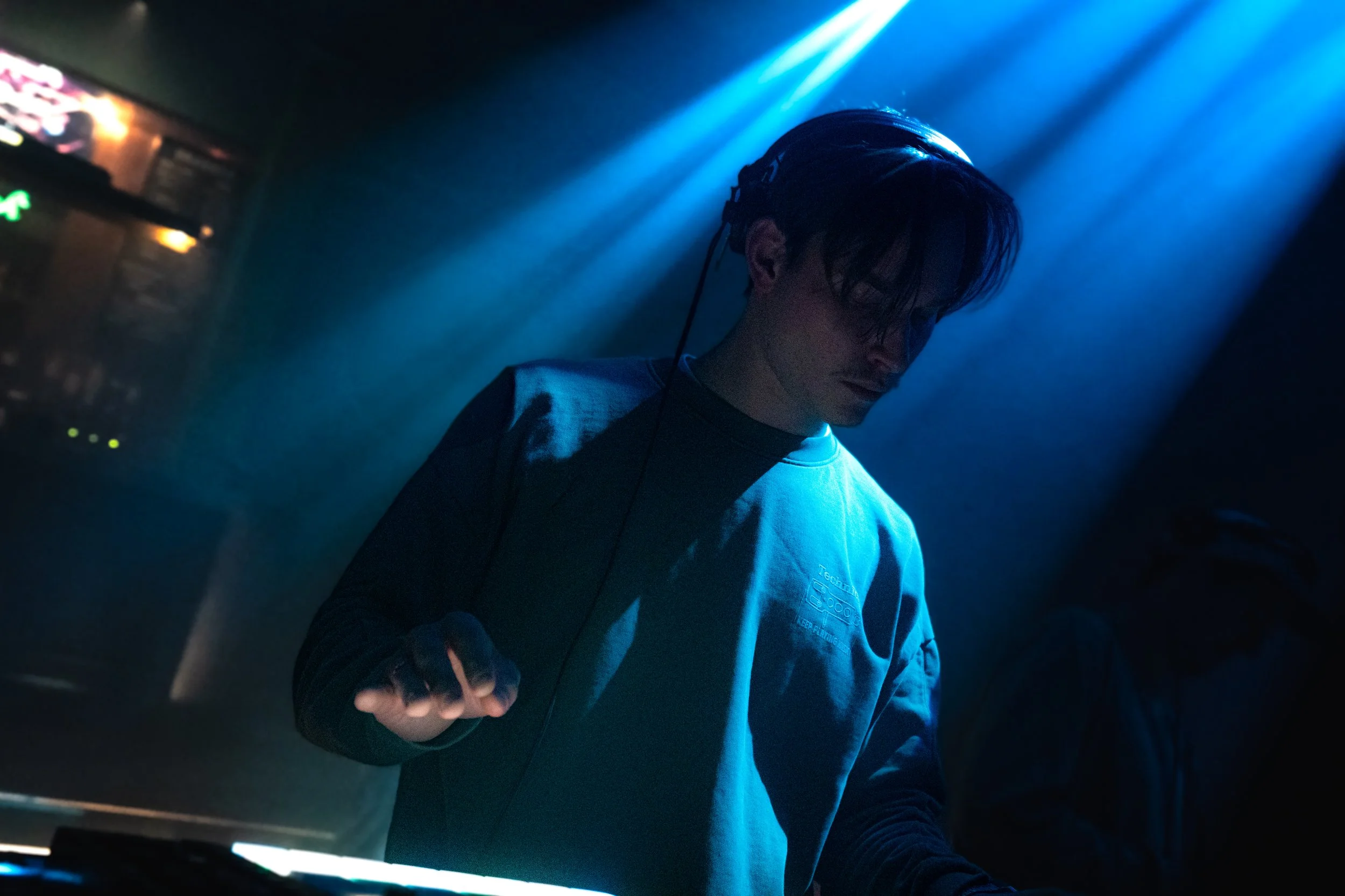 A person with dark hair wearing headphones and a light gray hoodie, standing in a dark room with blue lighting, possibly a nightclub or music venue.