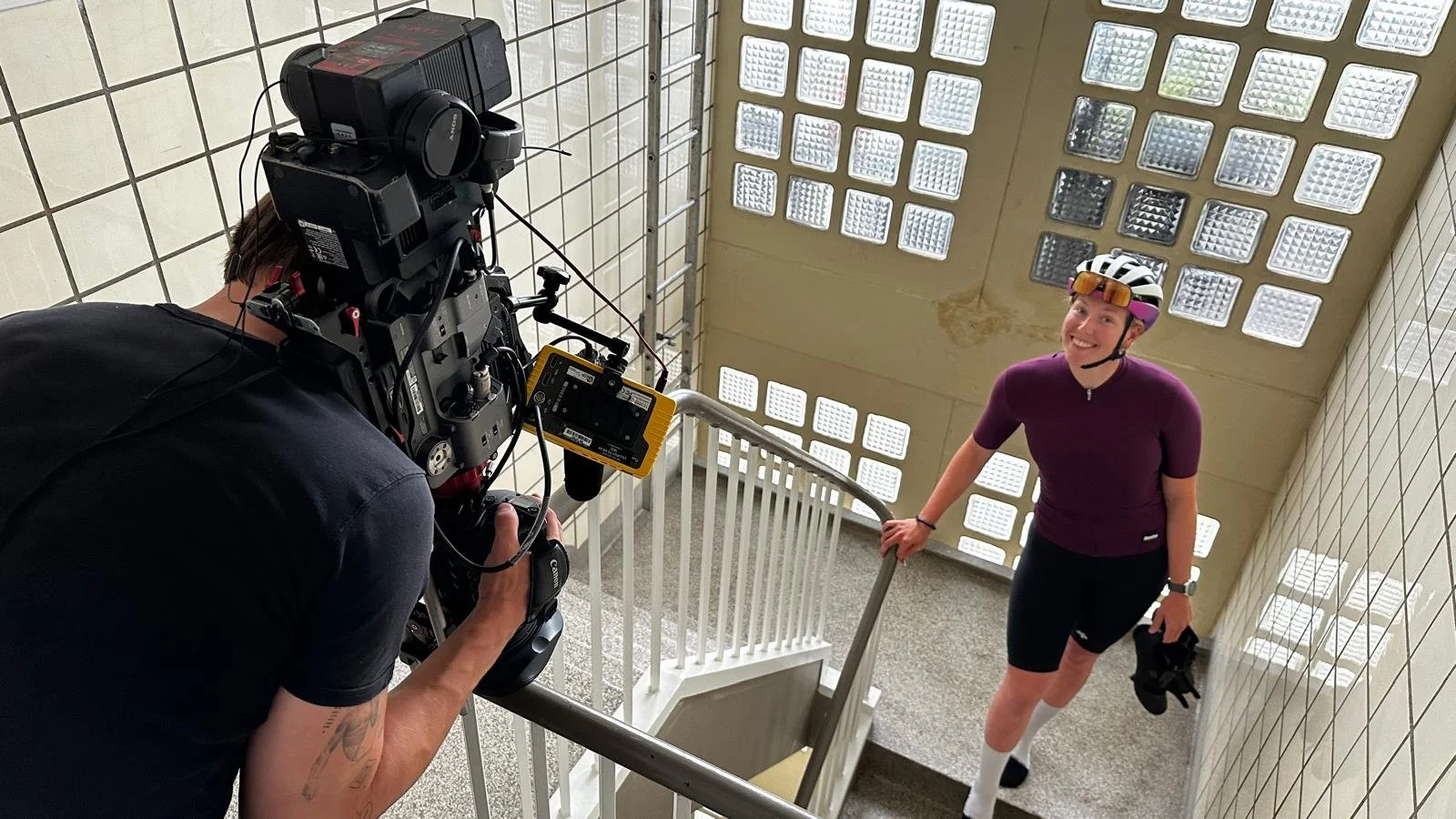 A woman in a purple cycling jersey, black shorts, and a helmet, smiling at the camera while holding cycling gloves, standing on a staircase landing with a tiled wall and glass block window behind her. A cameraman in black is taking her picture from above.