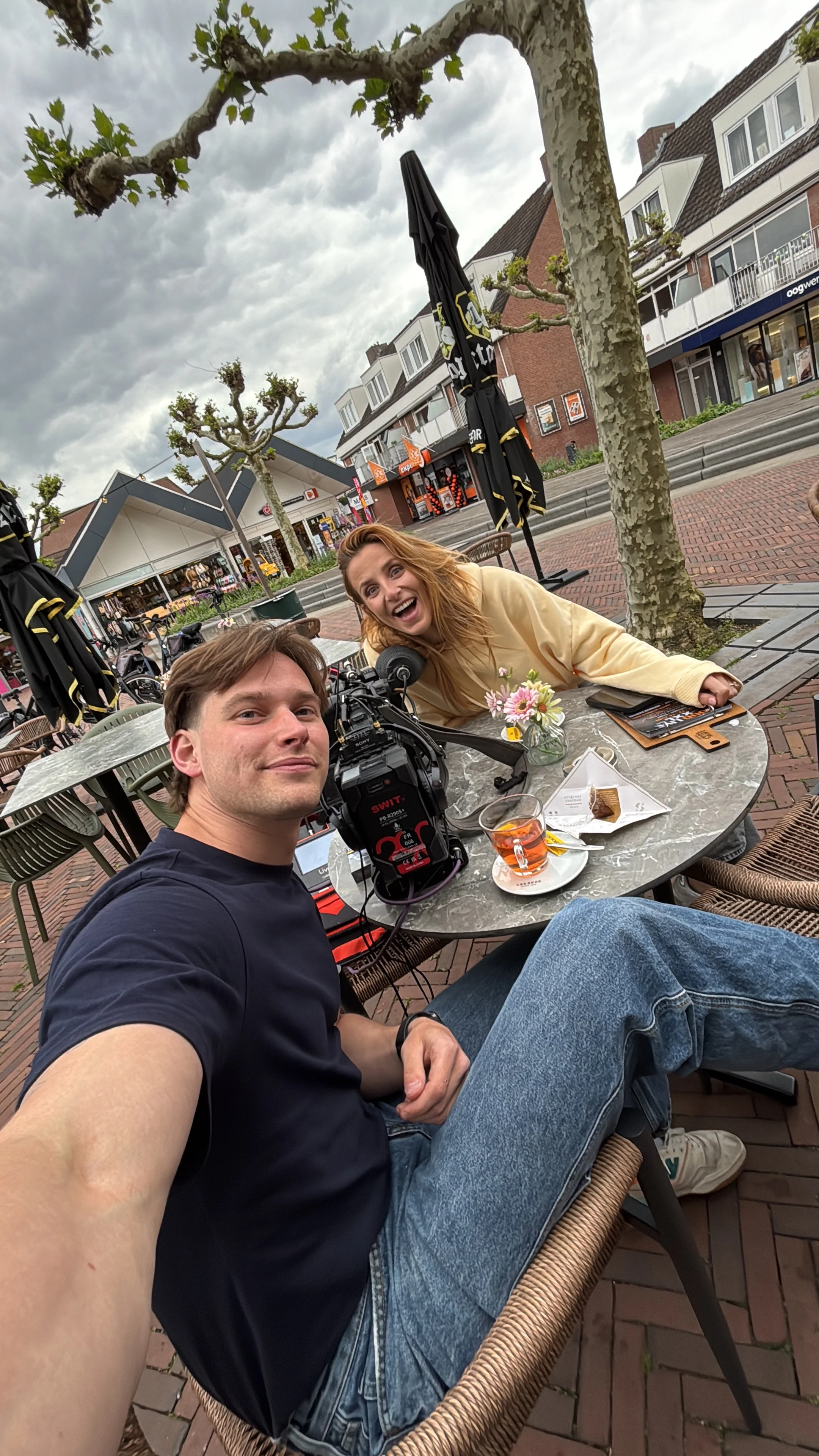 Two people sitting at a round outdoor table with food and drinks, taking a selfie. Behind them are leafless trees, buildings, and shops in a pedestrian area with a cloudy sky.