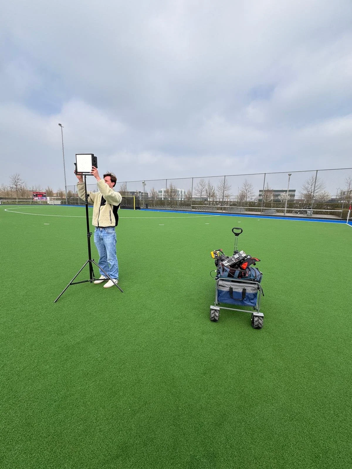 Person standing on a synthetic sports field adjusting lighting equipment, with tools and a cart nearby.