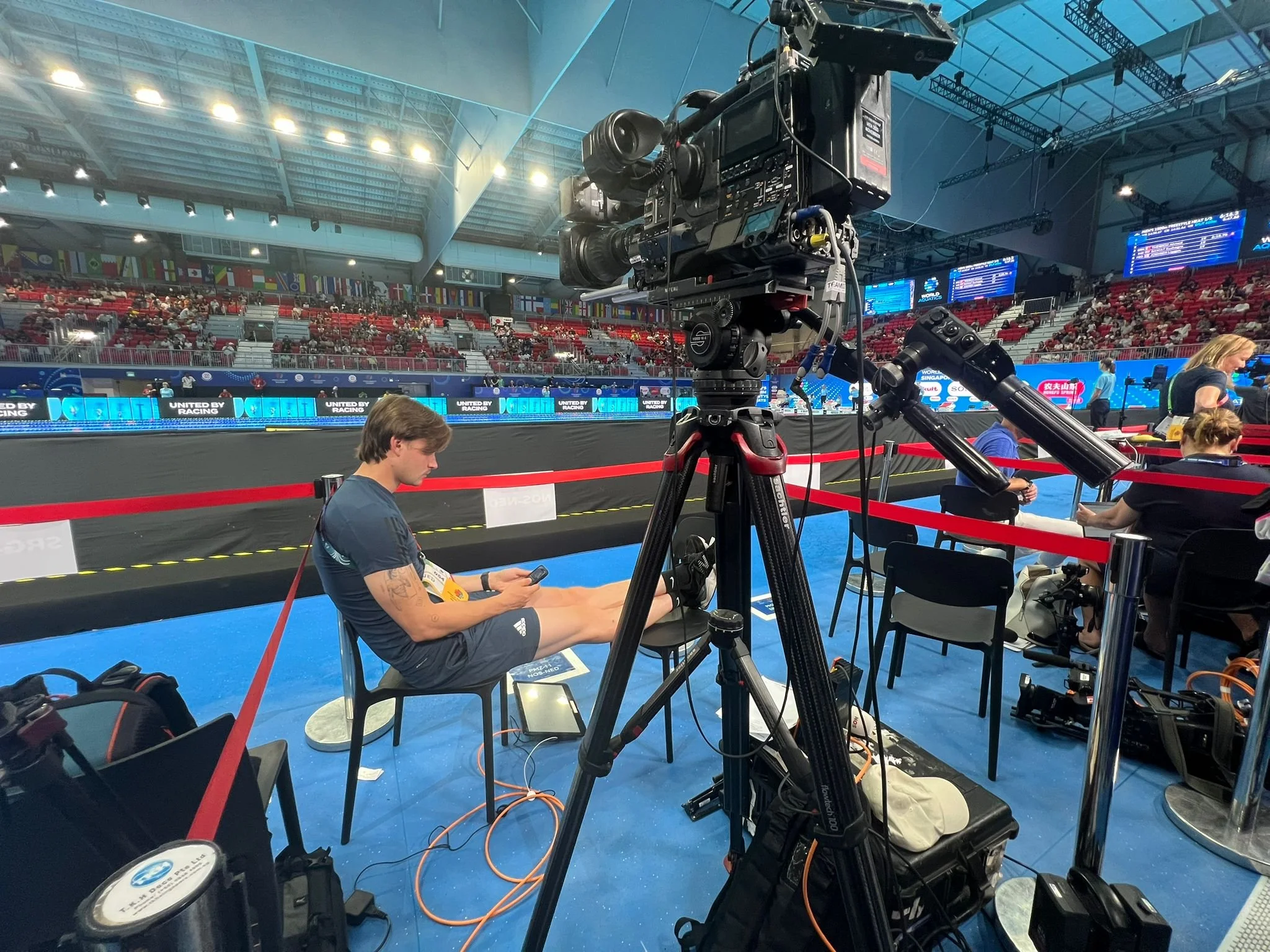 Inside an indoor sports arena with a large professional video camera on a tripod focused on the field. Several people are seated around, some using phones, with a backdrop of empty red and gray seats and digital screens showing event information.