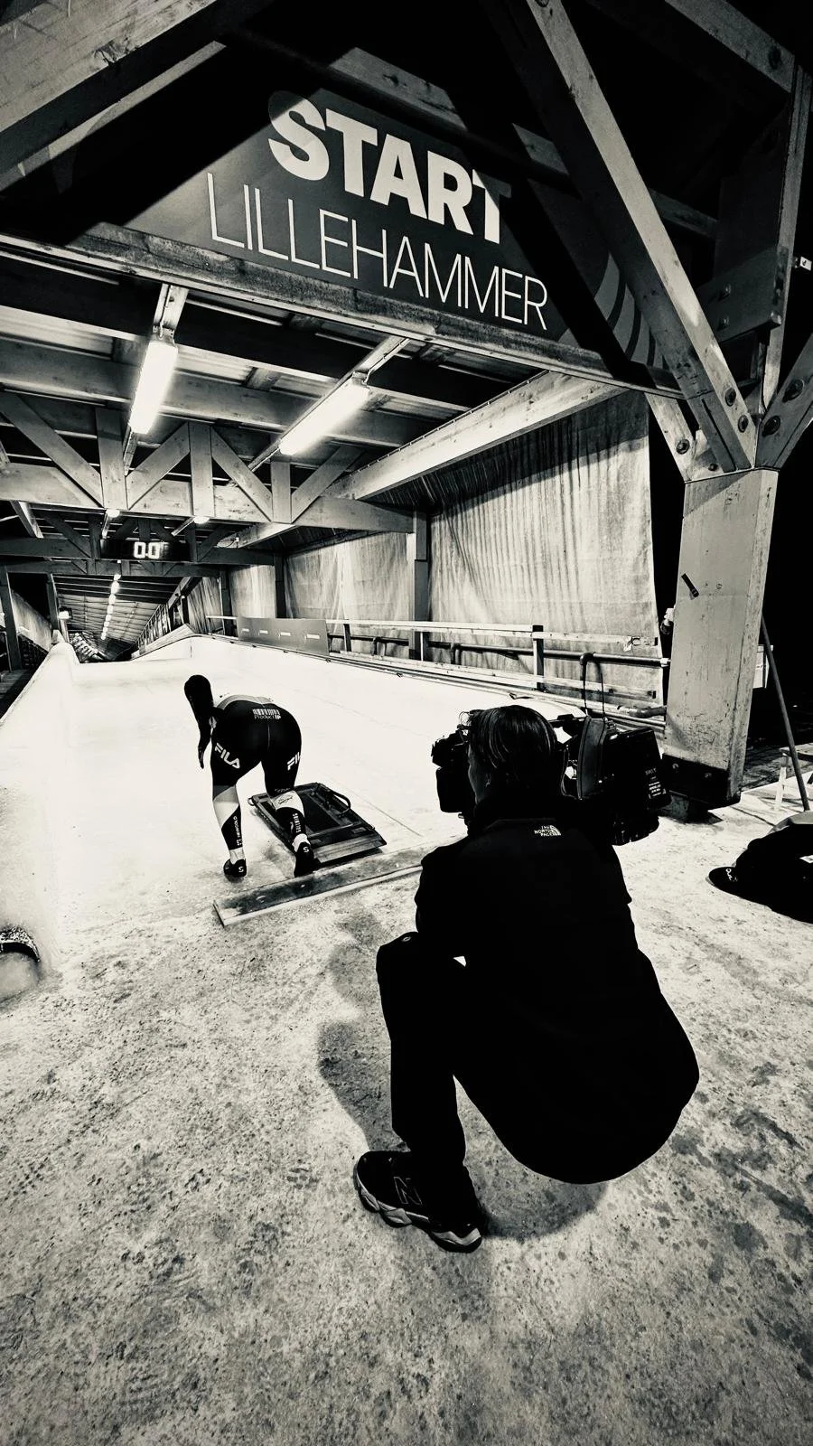 A person in athletic gear is preparing on an indoor track to slide down an ice track. A camera operator is kneeling nearby, filming the scene. The place is an Olympic bobsled track named Lillehammer, marked by a large sign overhead.