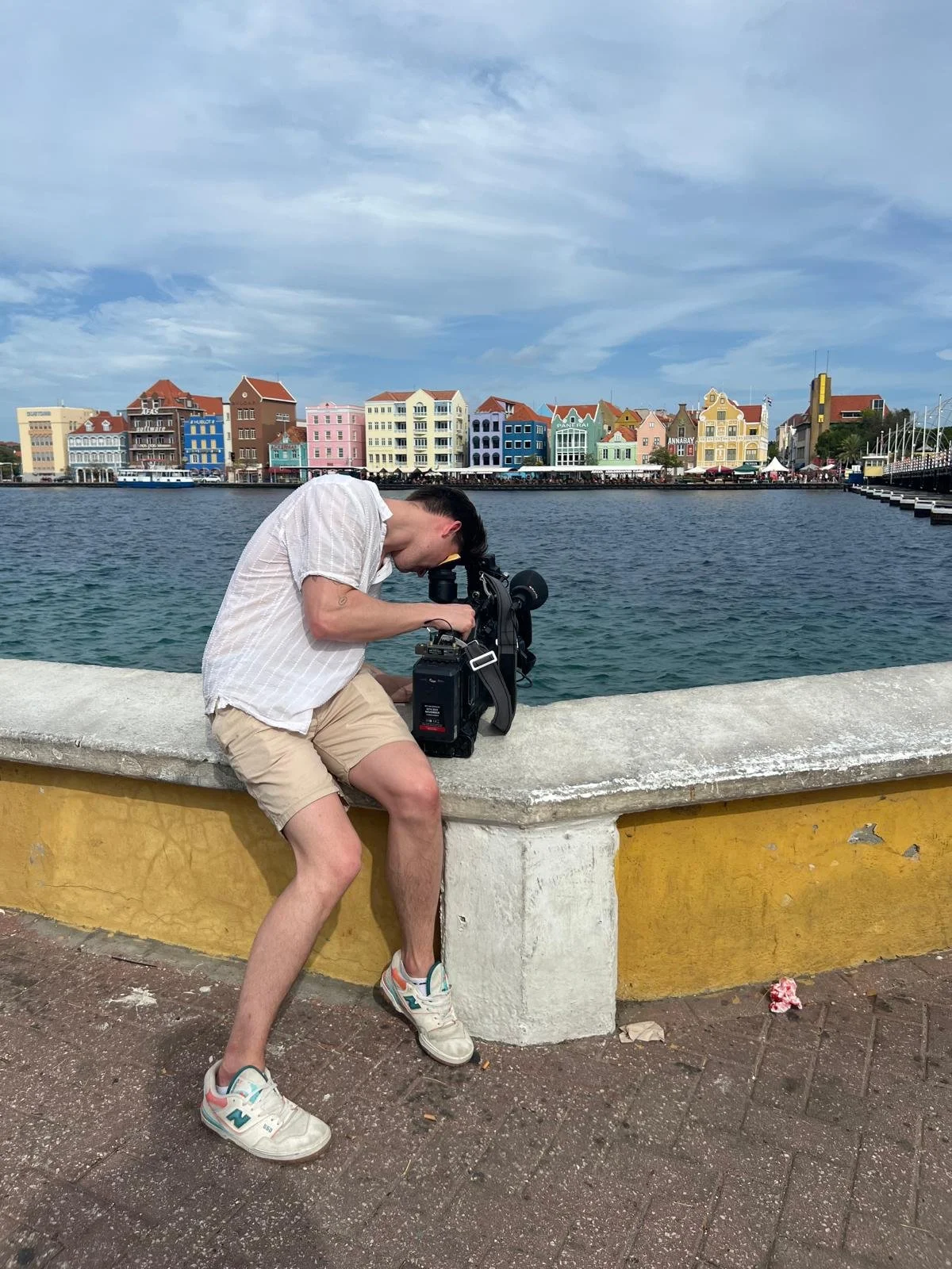 A man in a white shirt and beige shorts is crouched down on a concrete ledge, looking through a camera on a tripod, with colorful buildings across a body of water in the background.