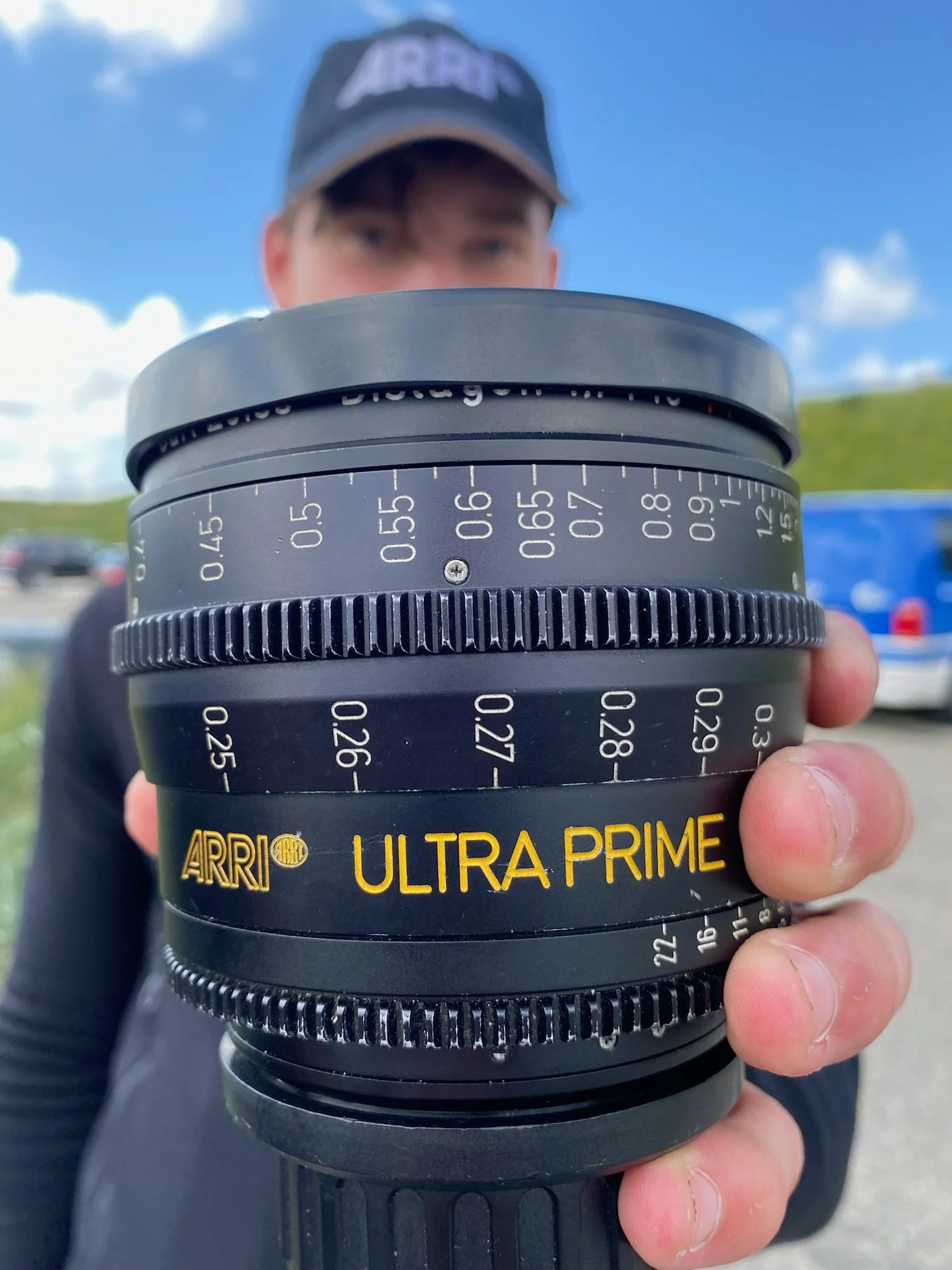 Close-up of a person holding a camera lens with a blue sky and clouds in the background. The person is wearing a cap and a dark jacket.