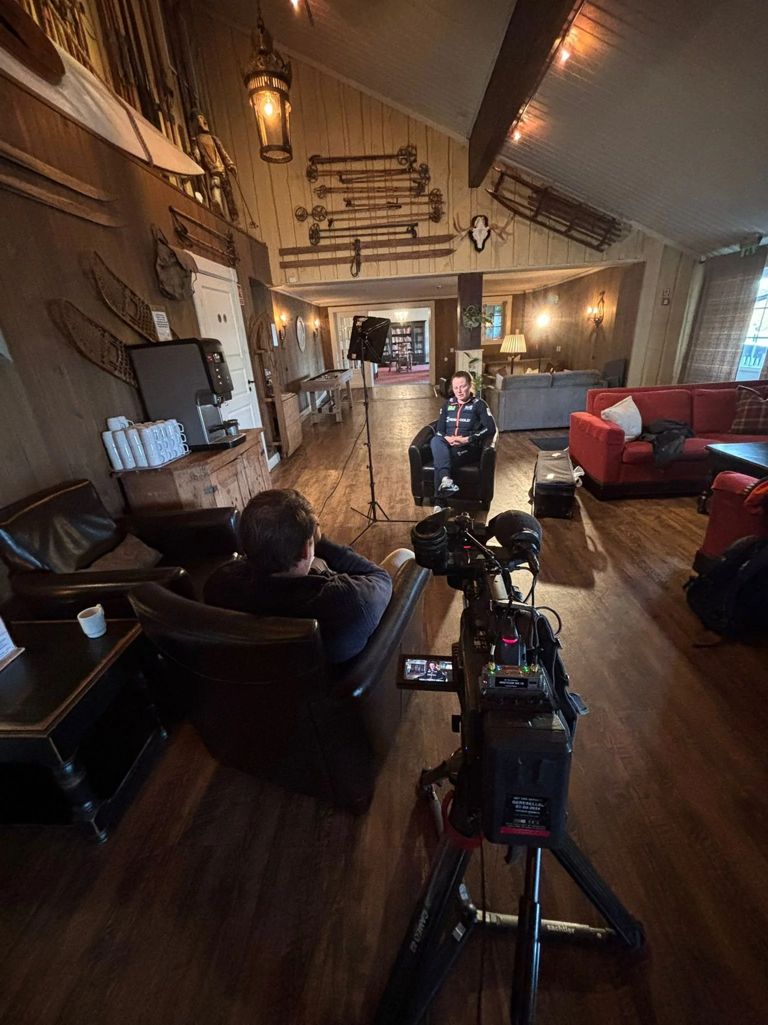 Interior of a rustic living room decorated with vintage tools, furniture, and hunting trophies. A woman is seated in front of a camera setup. The room includes wood-paneled walls, couches, a coffee table, and dim lighting.
