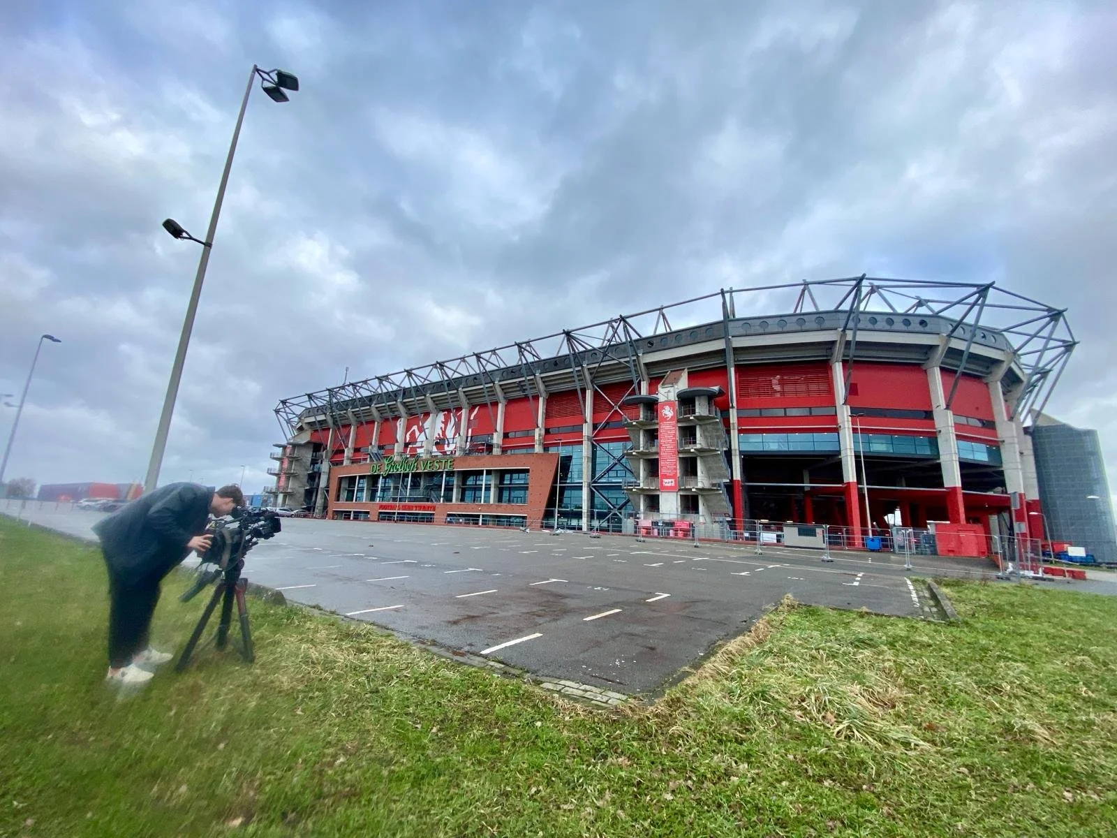 A person setting up or adjusting a camera on a tripod outside a large stadium with red and gray exterior, curved roof, and seating structure visible. The parking lot in front has no cars parked.