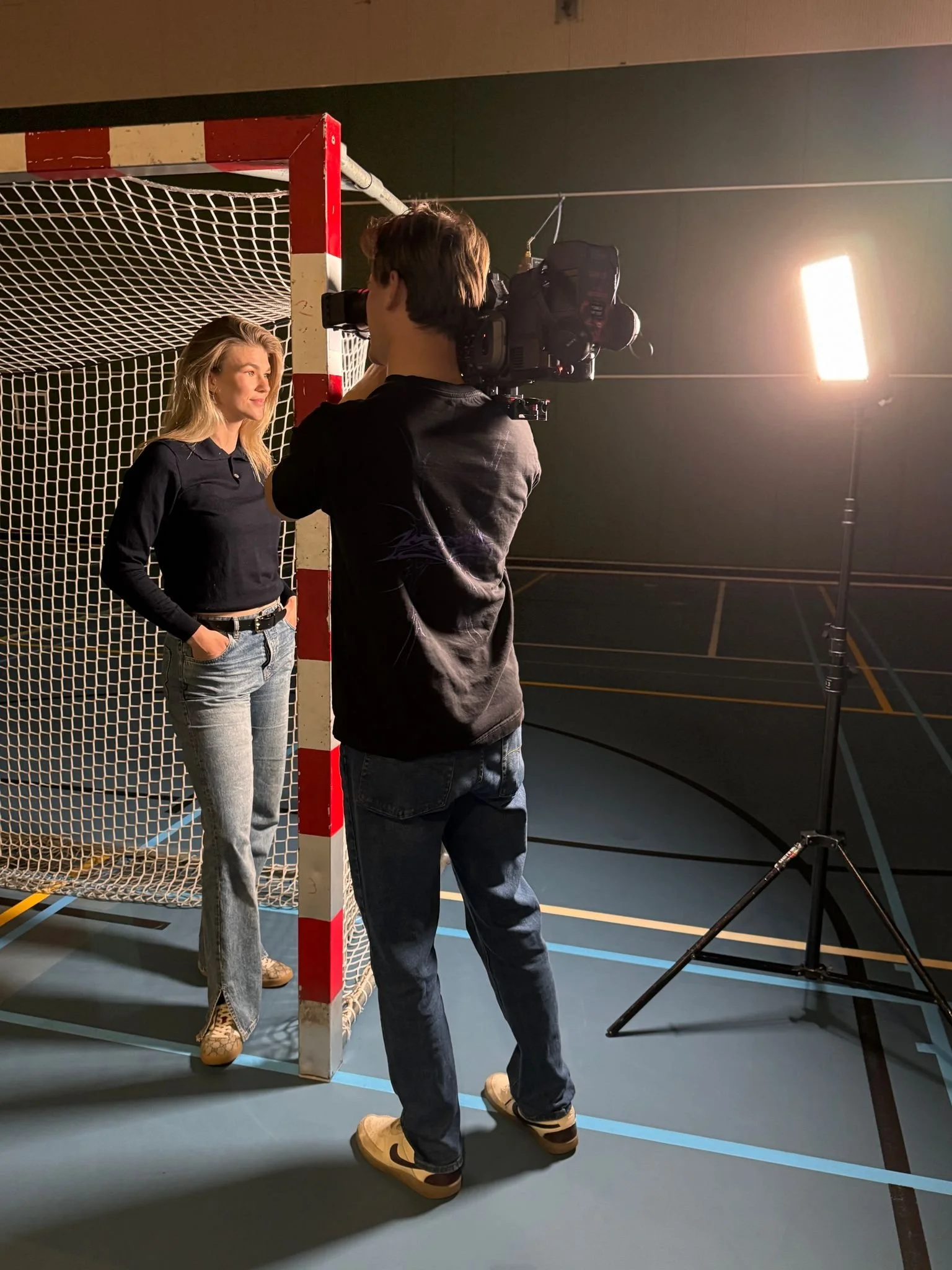 A woman poses for a photoshoot in front of a soccer goal on an indoor court, while a man takes her picture with a professional camera. The scene is lit with a large studio light.