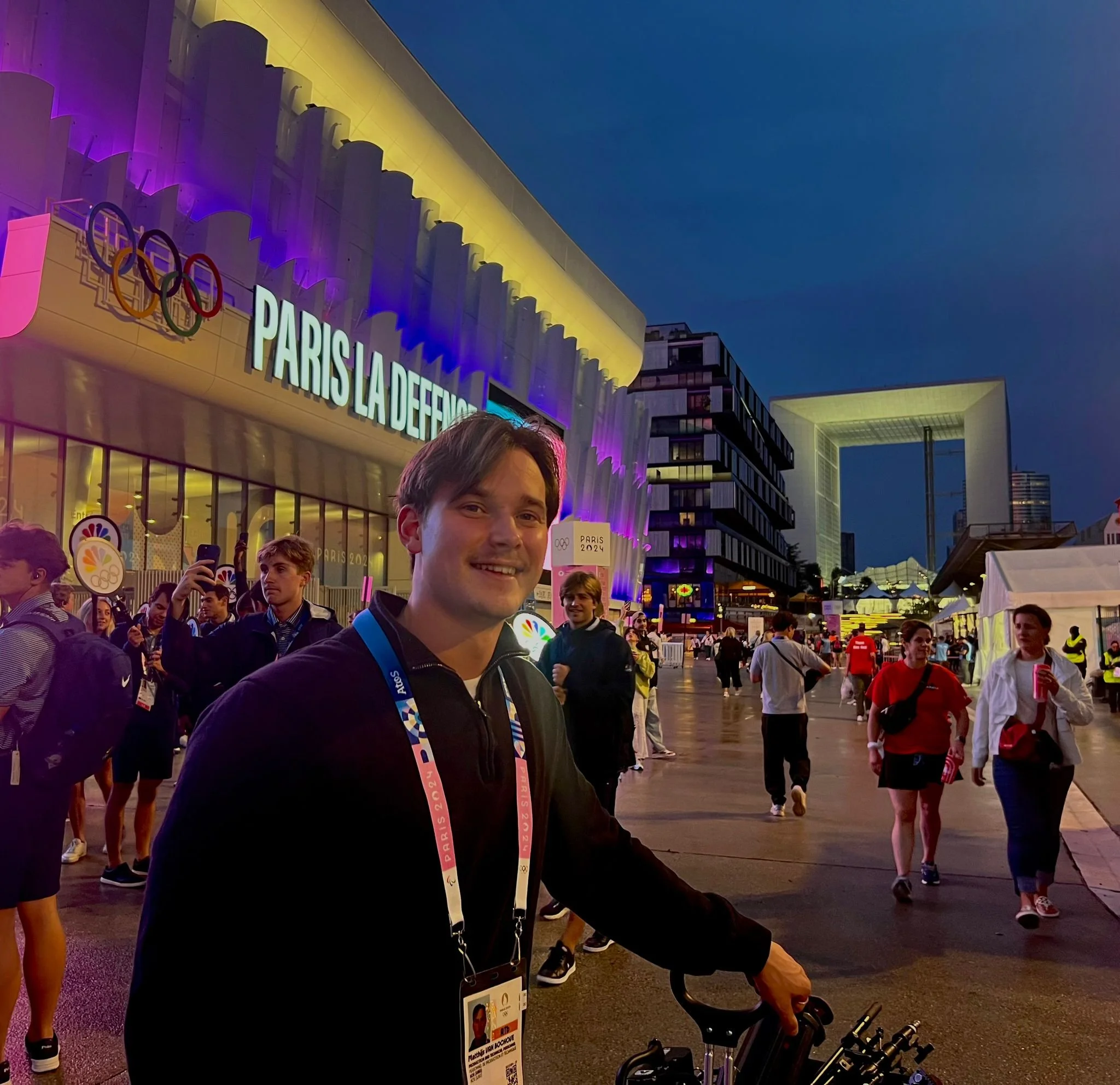 A smiling person in black clothing with a lanyard and badge around their neck stands outdoors at an Olympic event during evening, with a crowd of people and buildings illuminated with colorful lights in the background.