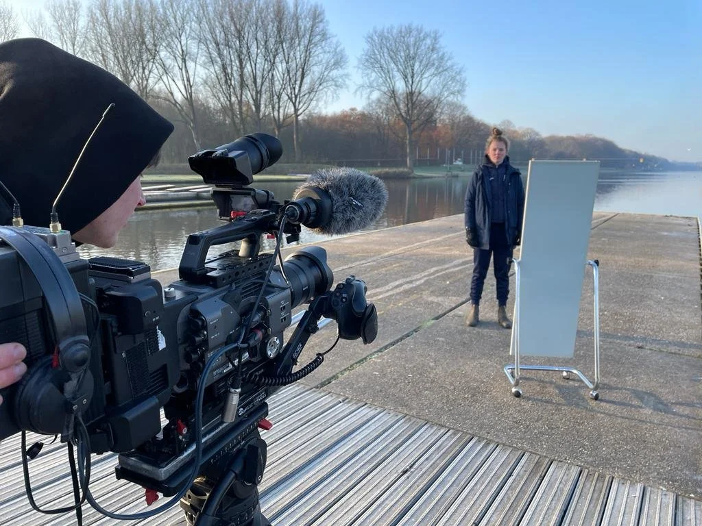 Person operating a video camera filming a woman standing outdoors near a body of water with a whiteboard on a stand.