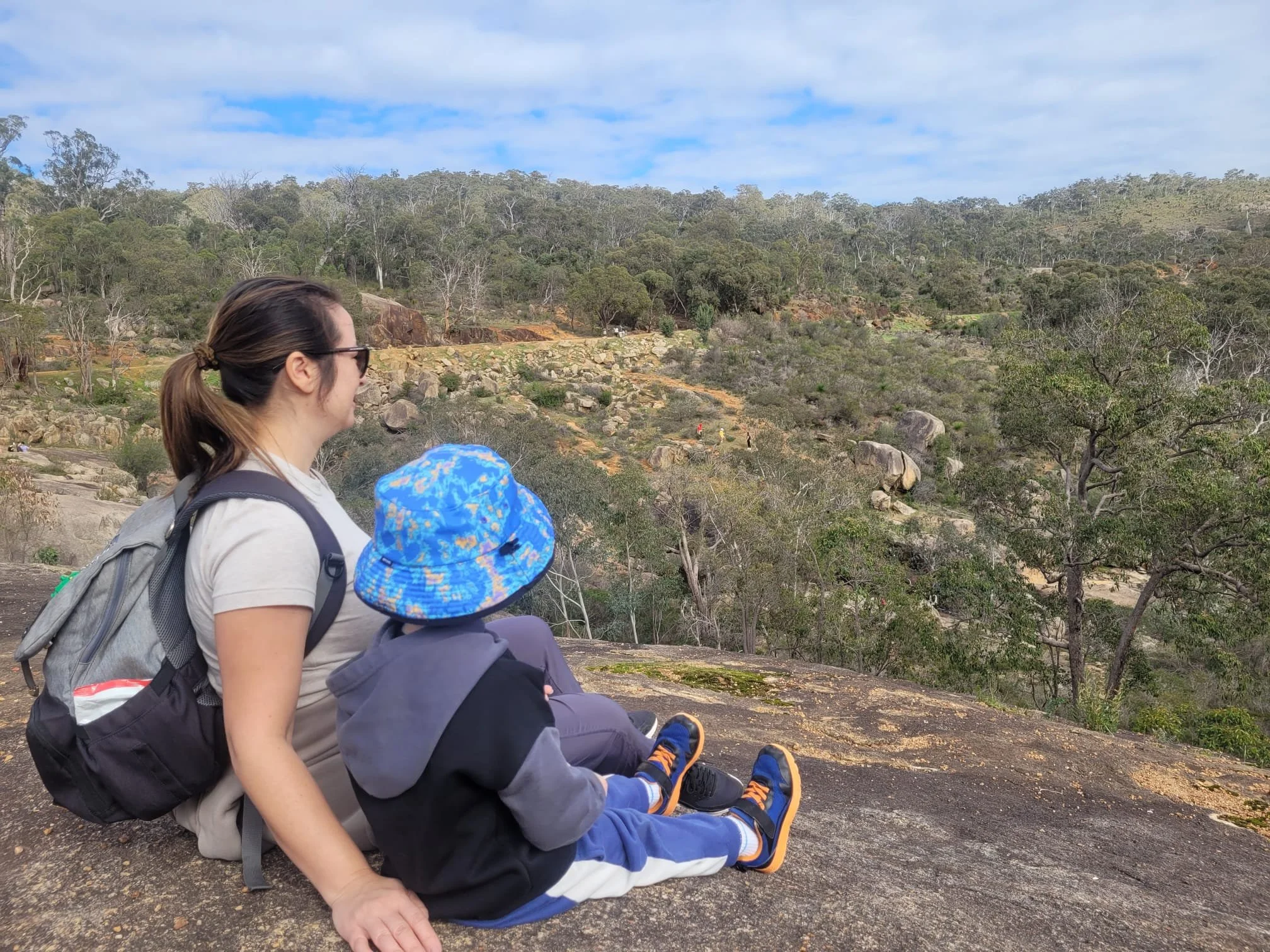 A woman and a child with backpacks sitting on the ground outdoors, overlooking a forested landscape with trees and rocks under a partly cloudy sky.