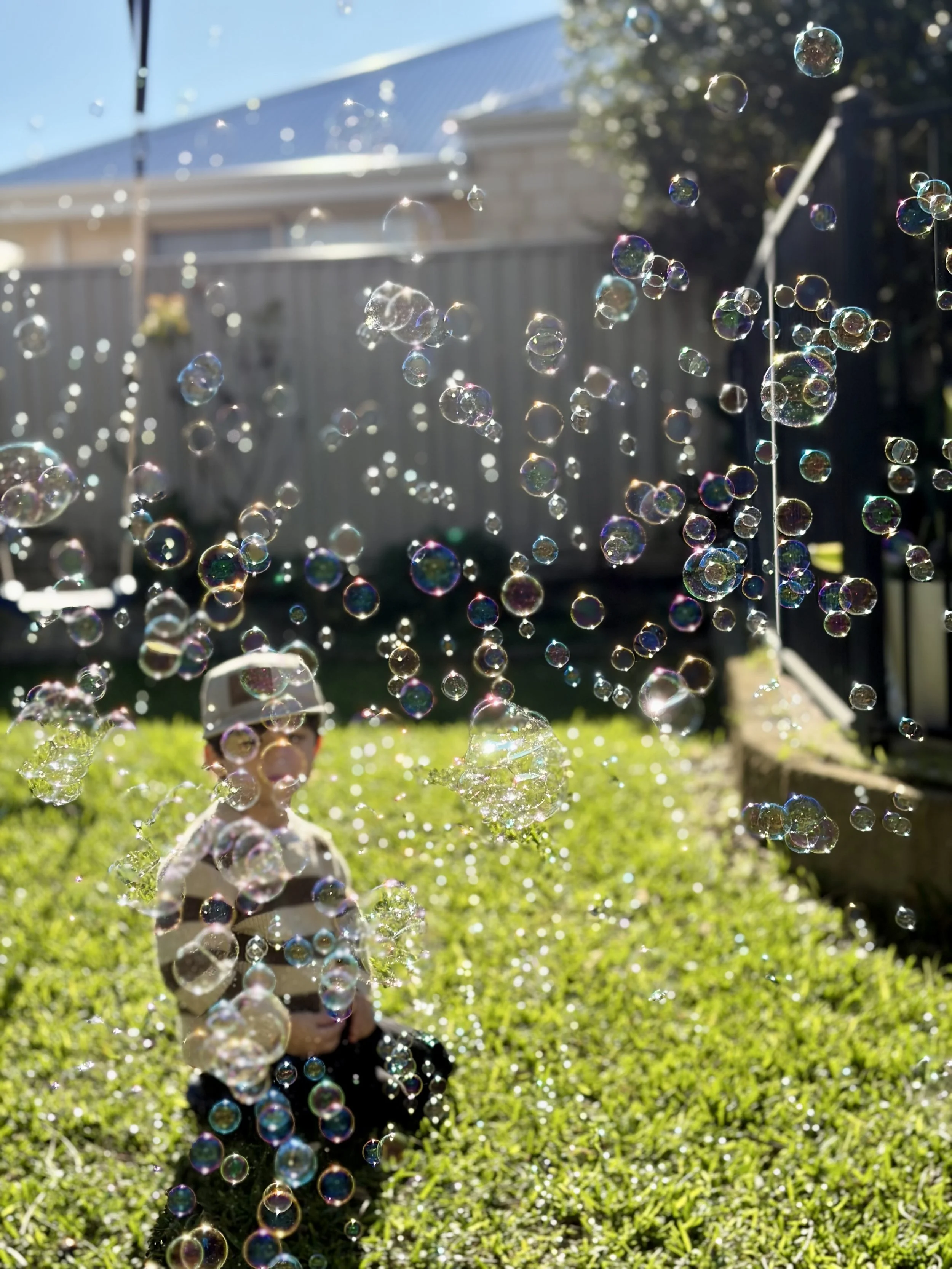 A young child wearing a white cap and black shirt is playing outside with soap bubbles during daytime, with a house and a fenced backyard visible in the background.