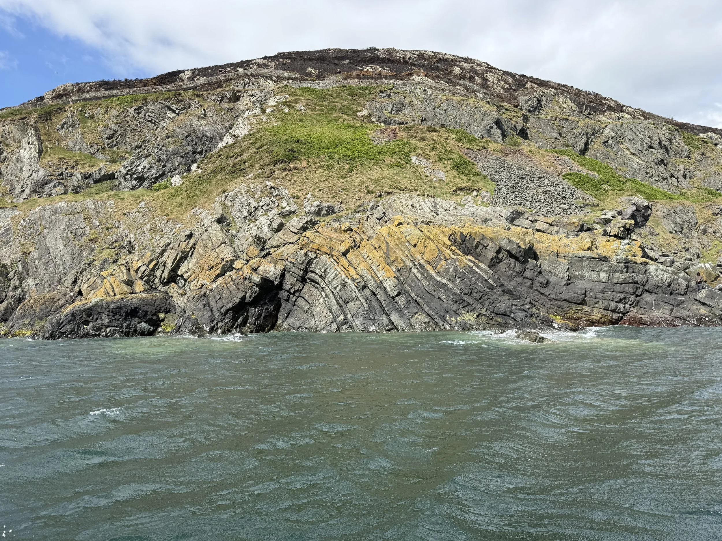 Cliffside landscape with layered rock formations and green vegetation, view from water level with blue sky and a few clouds.