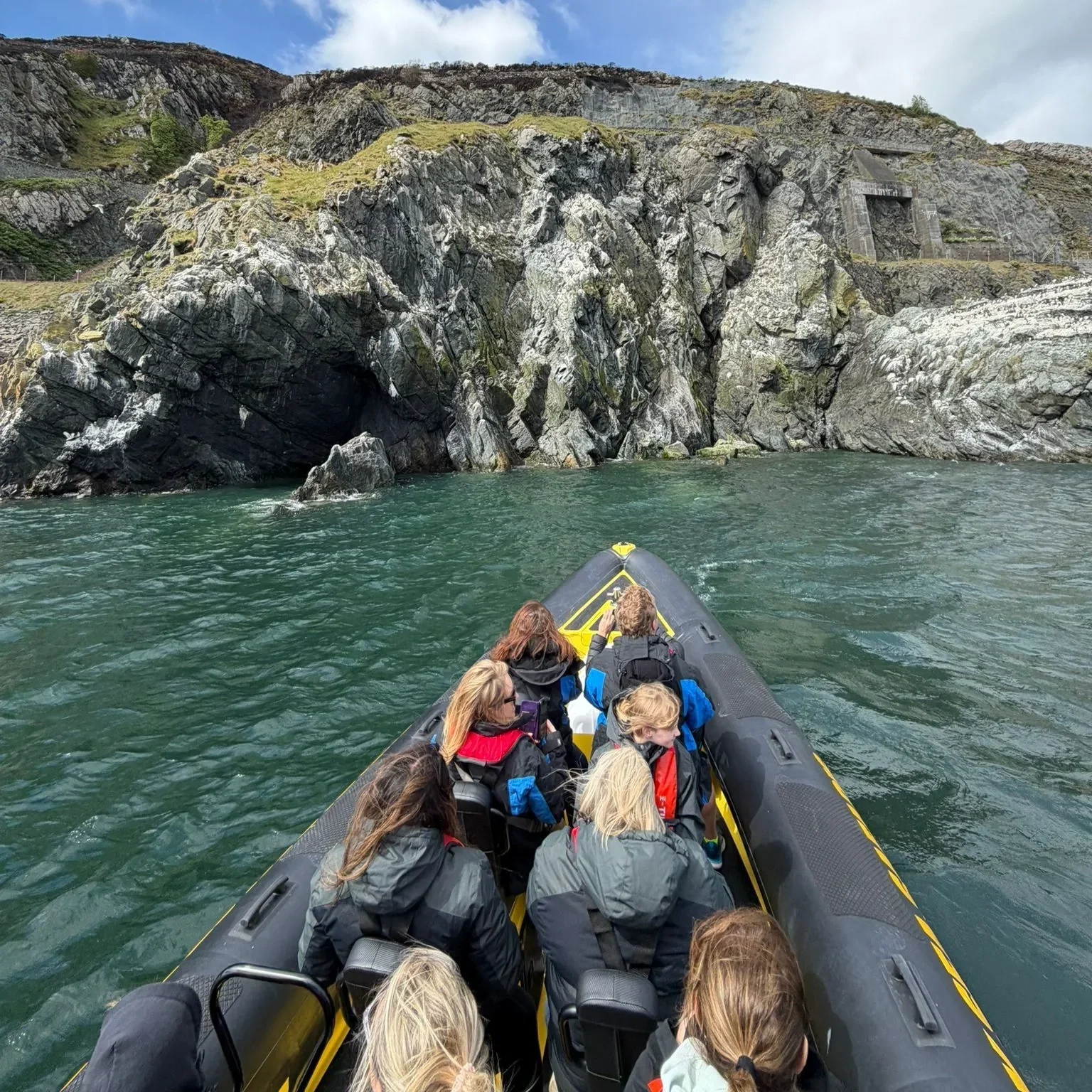 A group of people in life jackets riding in an inflatable boat on a body of water, approaching a large rocky cliff with stairs leading to a tunnel entrance.