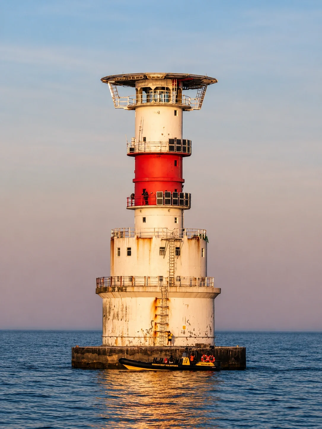 A tall, old lighthouse with red and white horizontal stripes standing in the water, with a boat nearby, under a clear blue sky.
