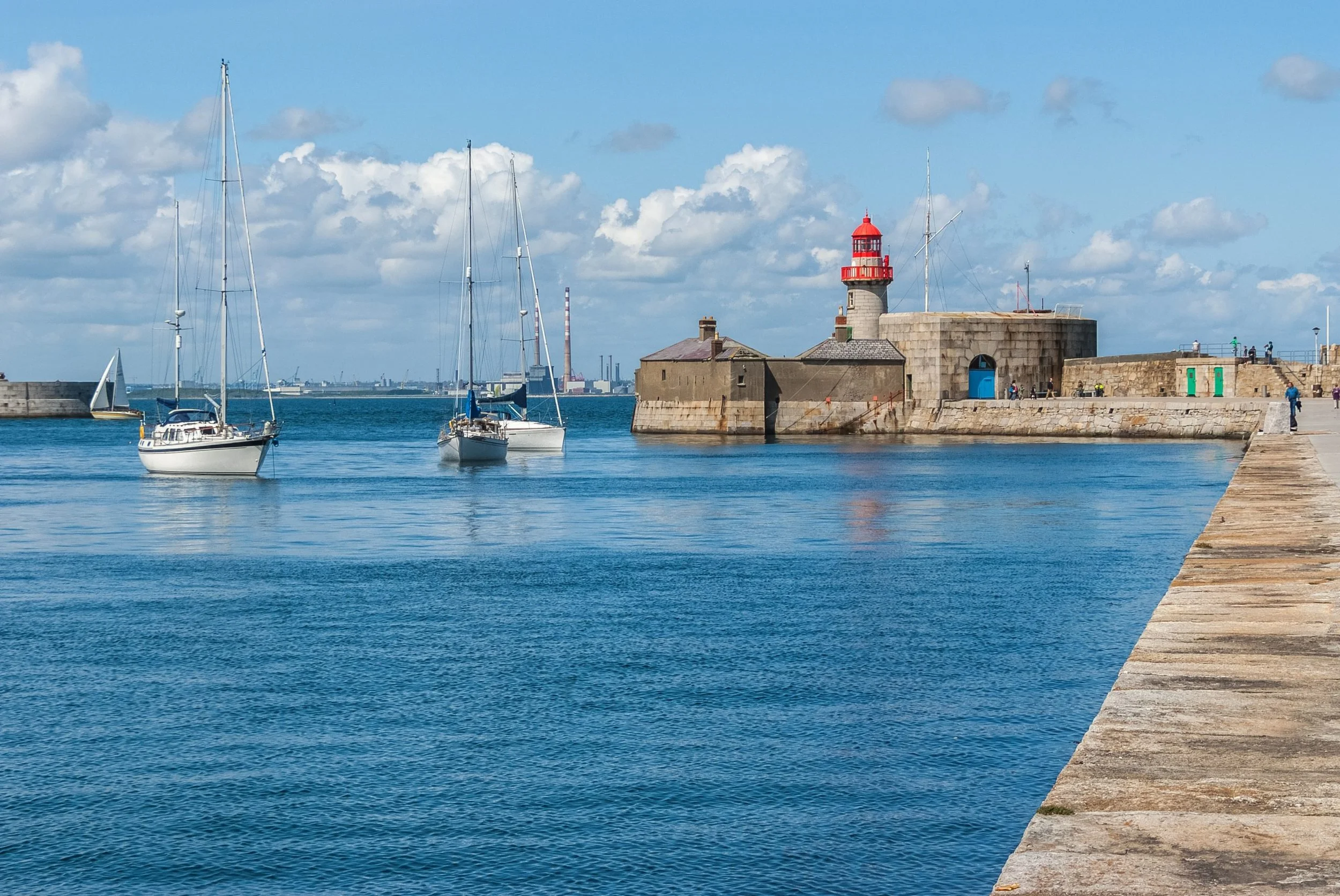 A harbor with sailing boats, a stone pier, and a lighthouse with a red top, under a partly cloudy sky.