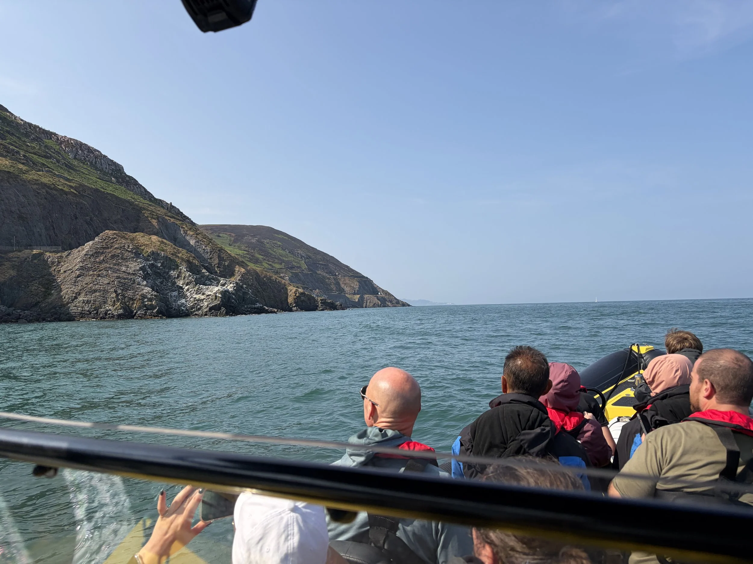 People on a boat ride near coastal cliffs on a sunny day, with the ocean and sailboat visible in the background.