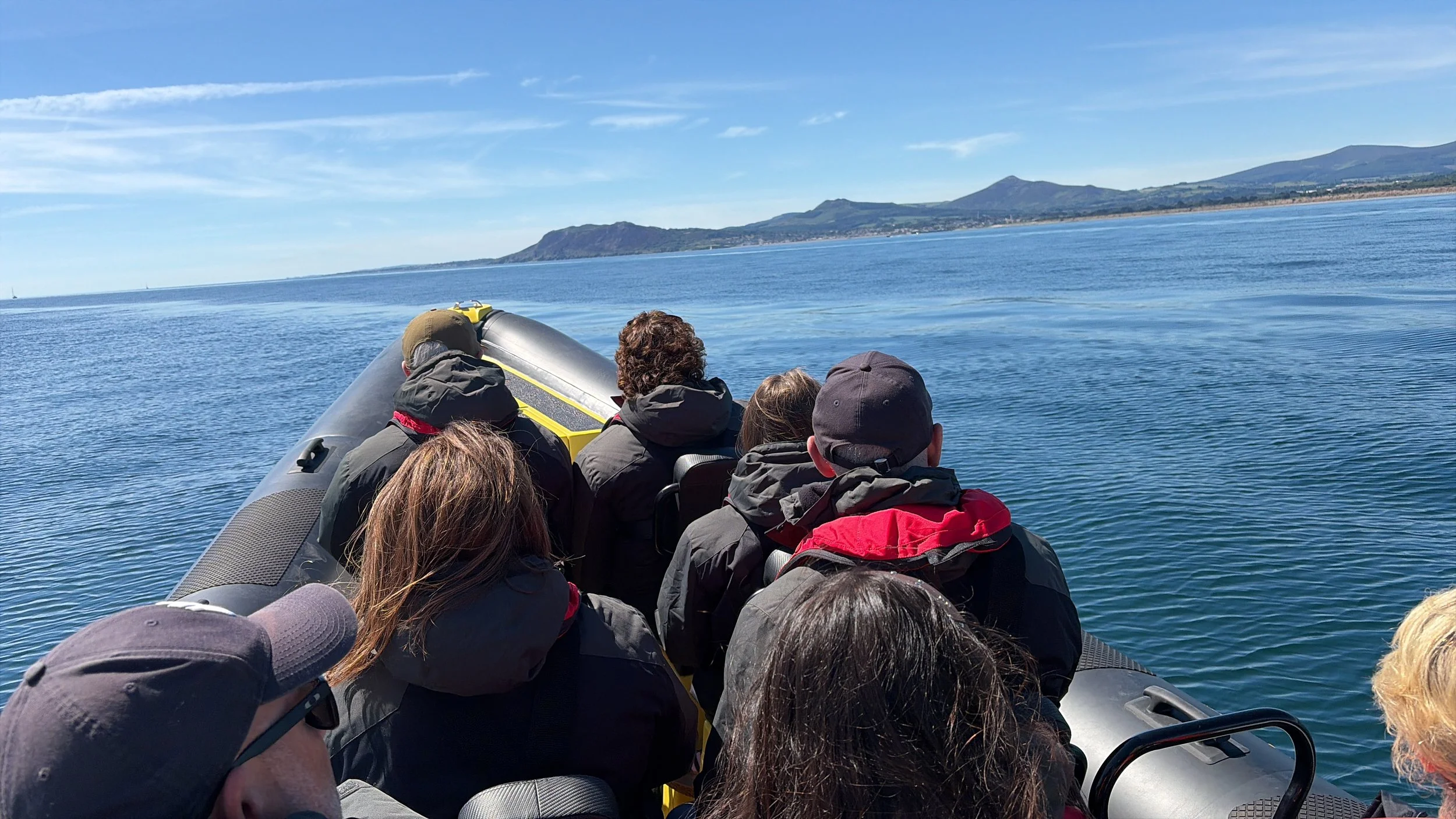 People sitting on a boat enjoying a ride on calm water with a scenic coastline and mountains in the background.