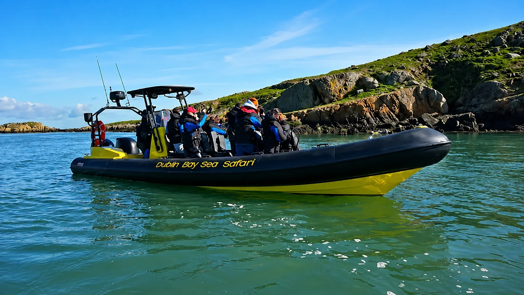 A group of people on a black and yellow boat labeled 'Dublin Bay Sea Safari' on a greenish water body, with rocky hills and a clear blue sky in the background.