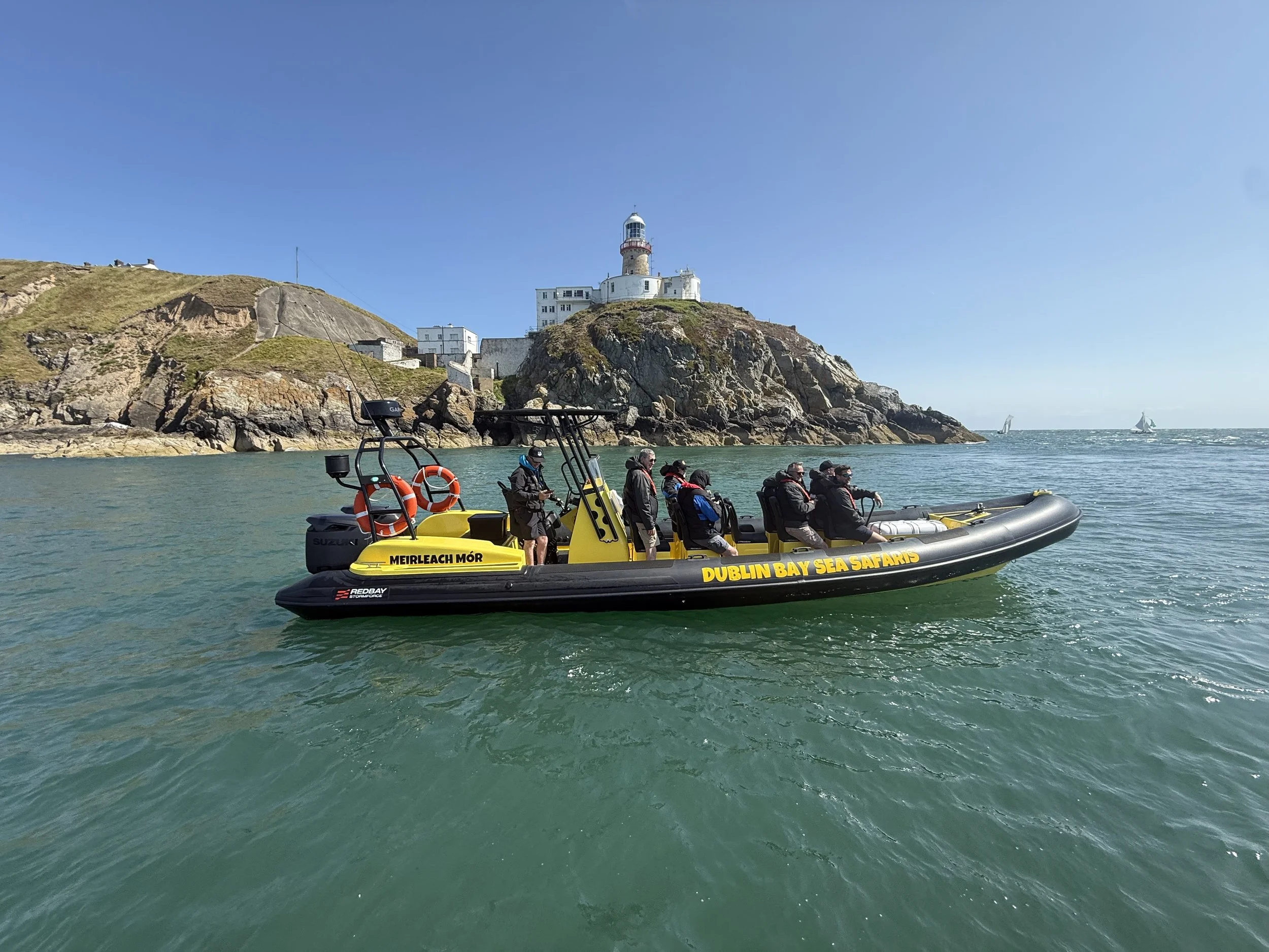 A group of people on a yellow boat with life rings, in Dublin Bay, sailing past a rocky coastal hill with a lighthouse on top at Ireland's Eye