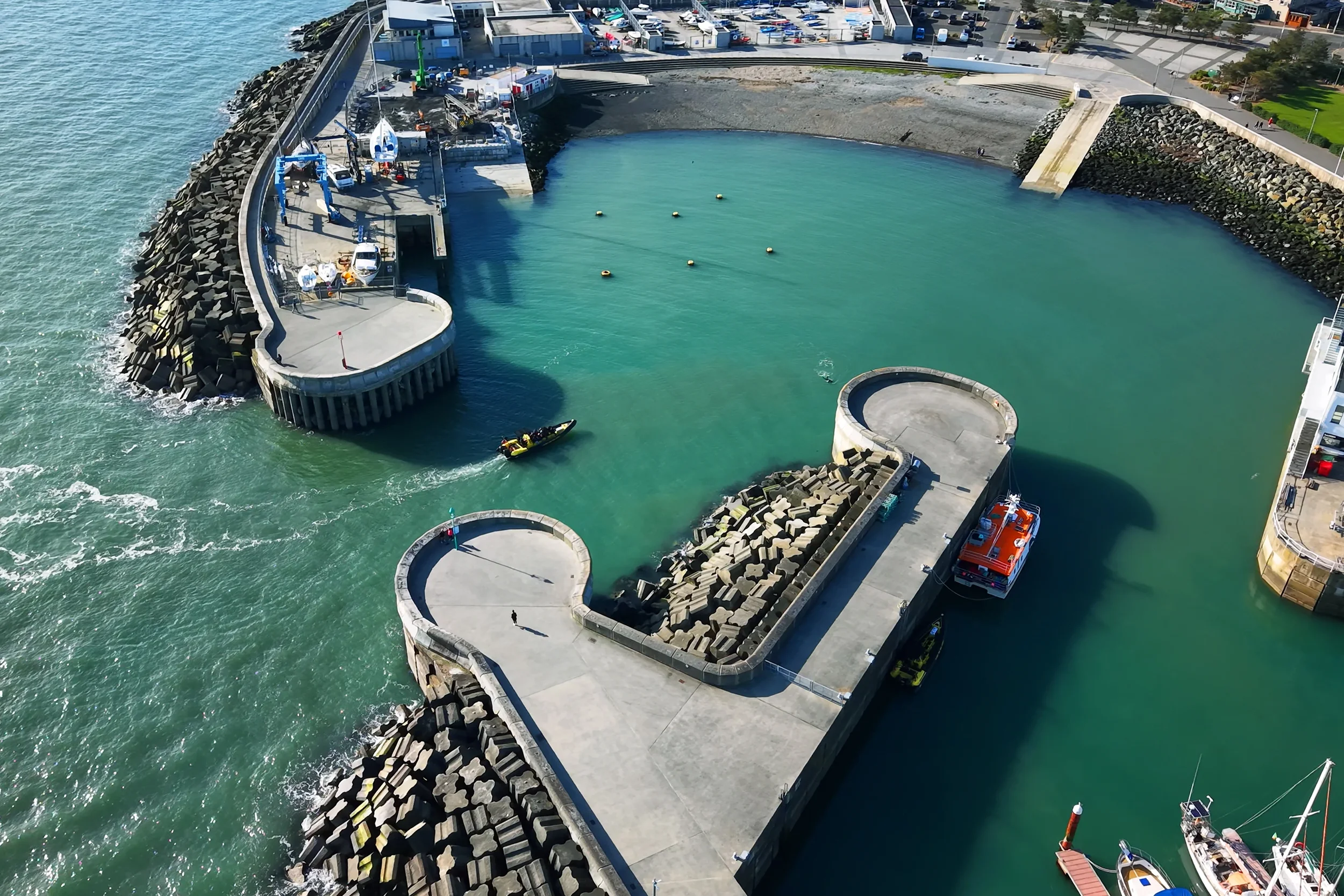 Aerial view of Greystones marina with boats and water, a breakwater with large rocks, and a parking lot with cars adjacent to a green park area.