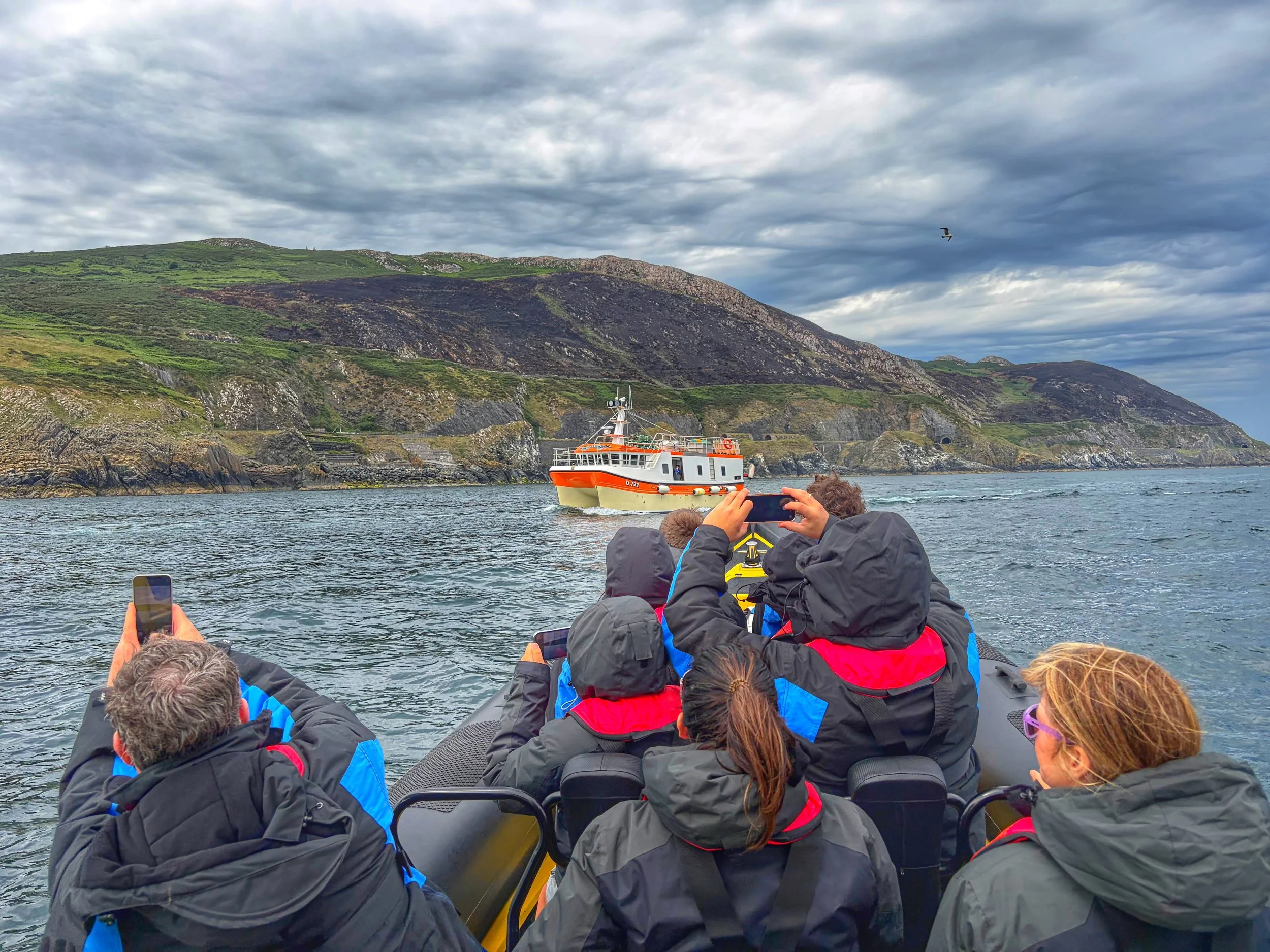 A RIB boat with multiple passengers wearing life jackets taking photos of Bray Head Co Wicklow with a fishing boat in the background