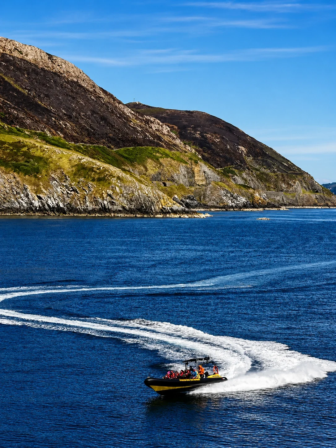 A speedboat with passengers speeding across the water near a rugged coastline with green hills and cliffs under a blue sky.