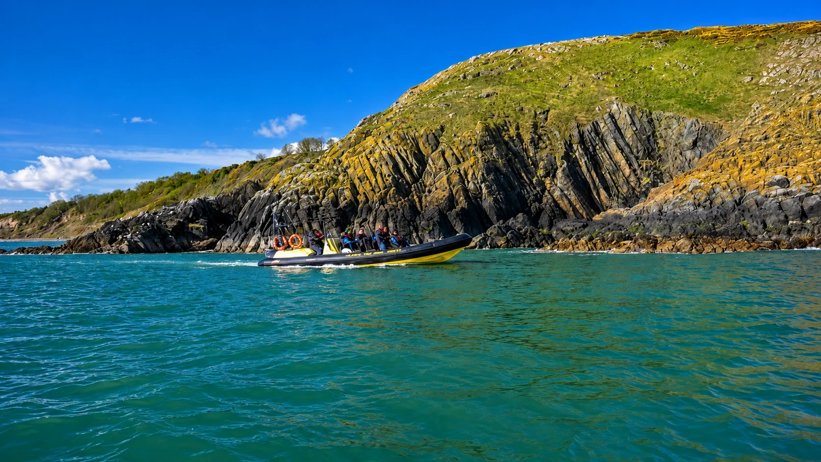 A group of people riding a yellow and black inflatable boat near a rocky cliff on a sunny day with blue sky and clear water.