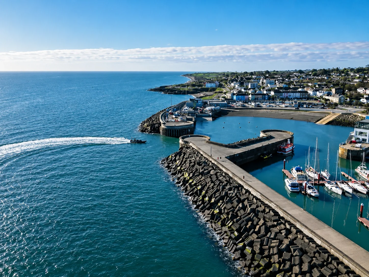 A harbor with boats docked and a breakwater extending into the ocean, with a town and green hills in the background under a partly cloudy sky.