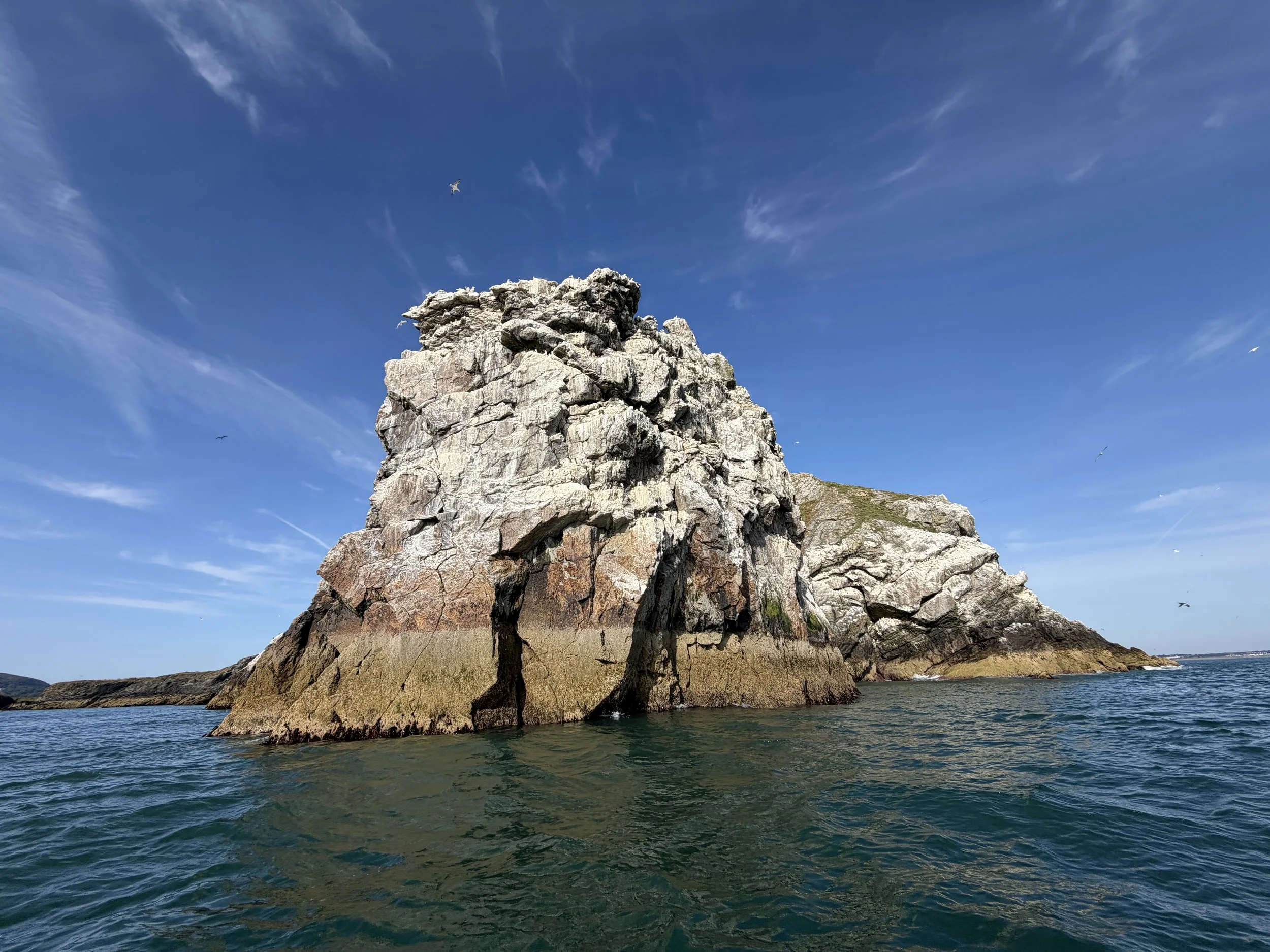 Large rocky island with steep cliffs surrounded by ocean under a blue sky with wispy clouds and a few birds flying.