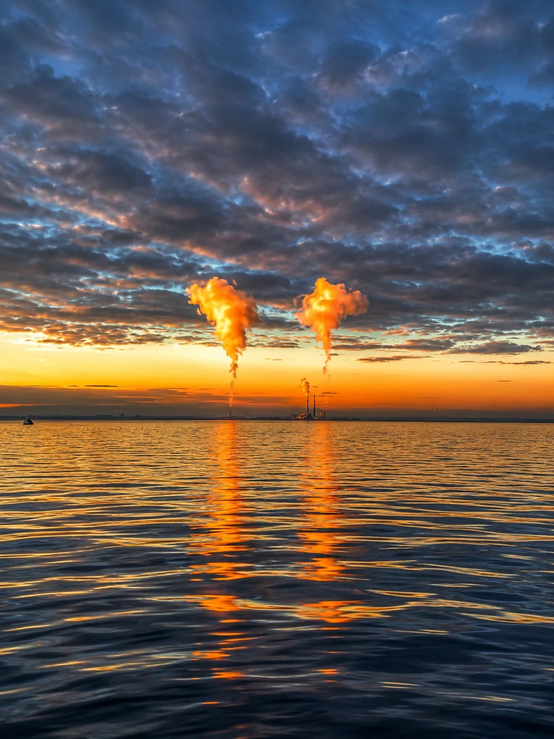 Sunset over water in Dublin Bay with industrial smoke stacks emitting orange smoke reflected on the water, with a partly cloudy sky.
