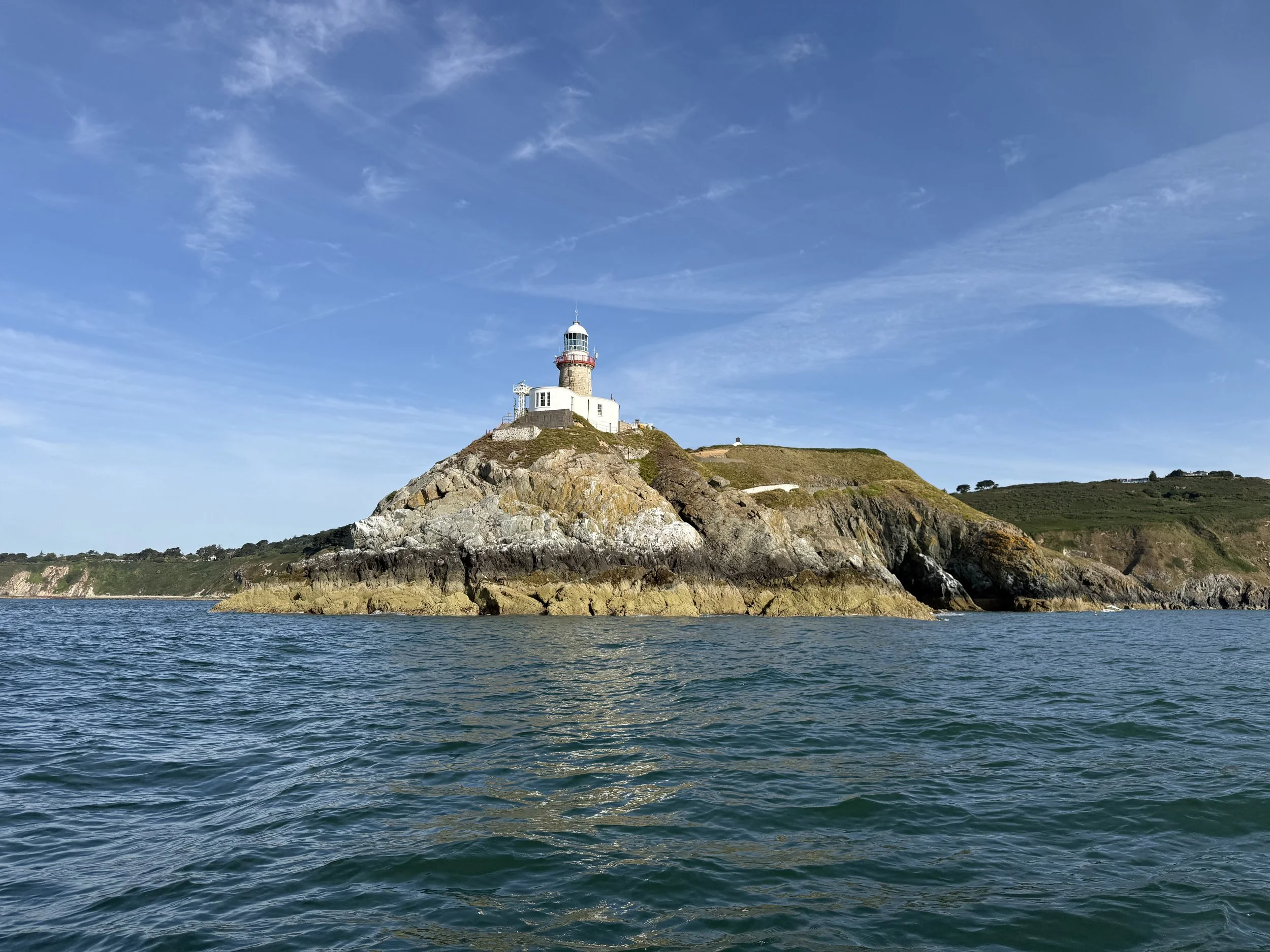 A lighthouse on a rocky hilltop surrounded by water, under a partly cloudy blue sky.