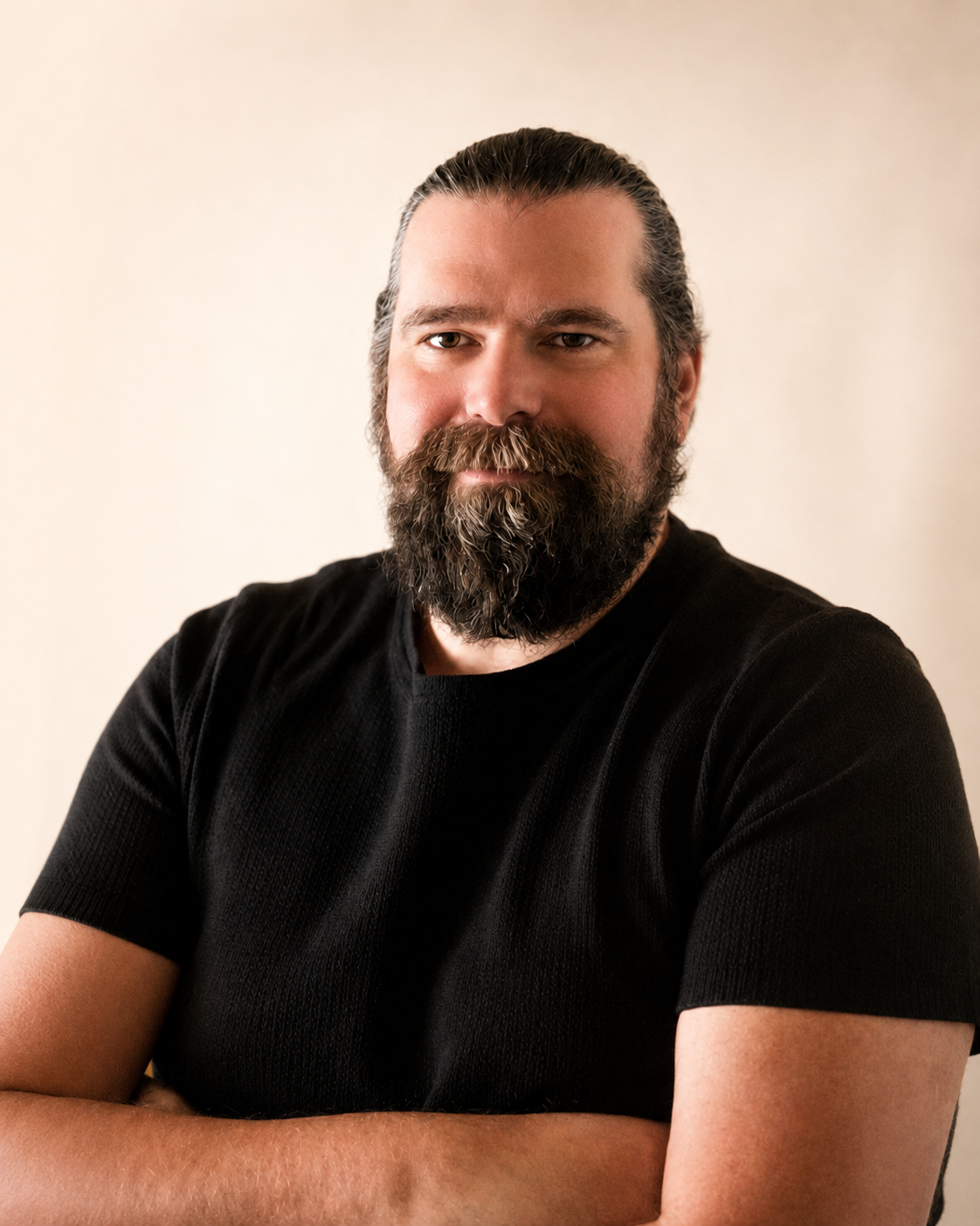 Un homme avec une barbe et une coiffure en arrière, portant un t-shirt noir, posant avec les bras croisés devant un fond neutre.