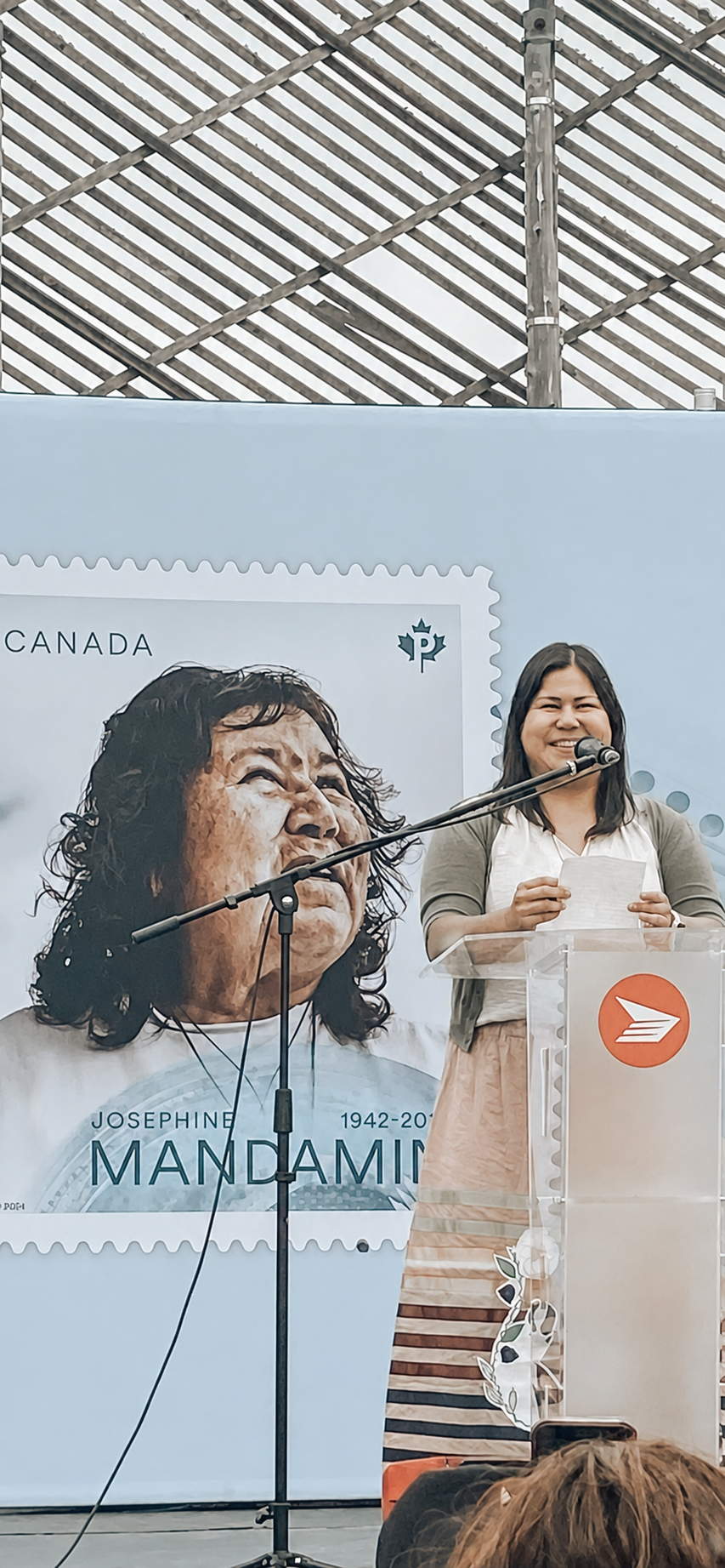 Regina Mandamin speaking at a podium during a tribute event for Josephine Mandamin in Canada, with a large poster of Josephine Mandamin's face in the background.
