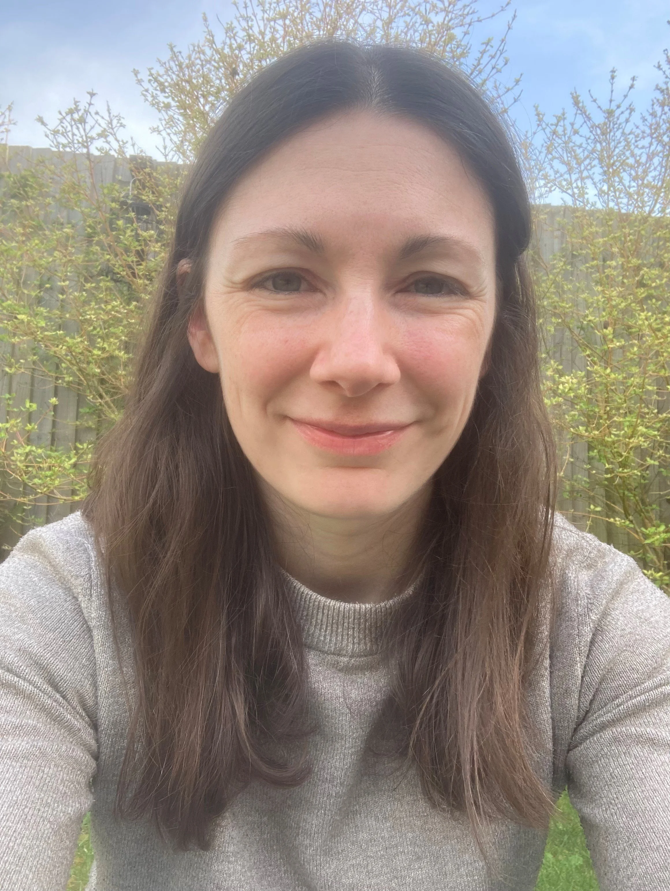 A woman with long brown hair and a light complexion smiling outdoors, wearing a grey jumper. She is in front of a fence with trees and blue sky in the background.