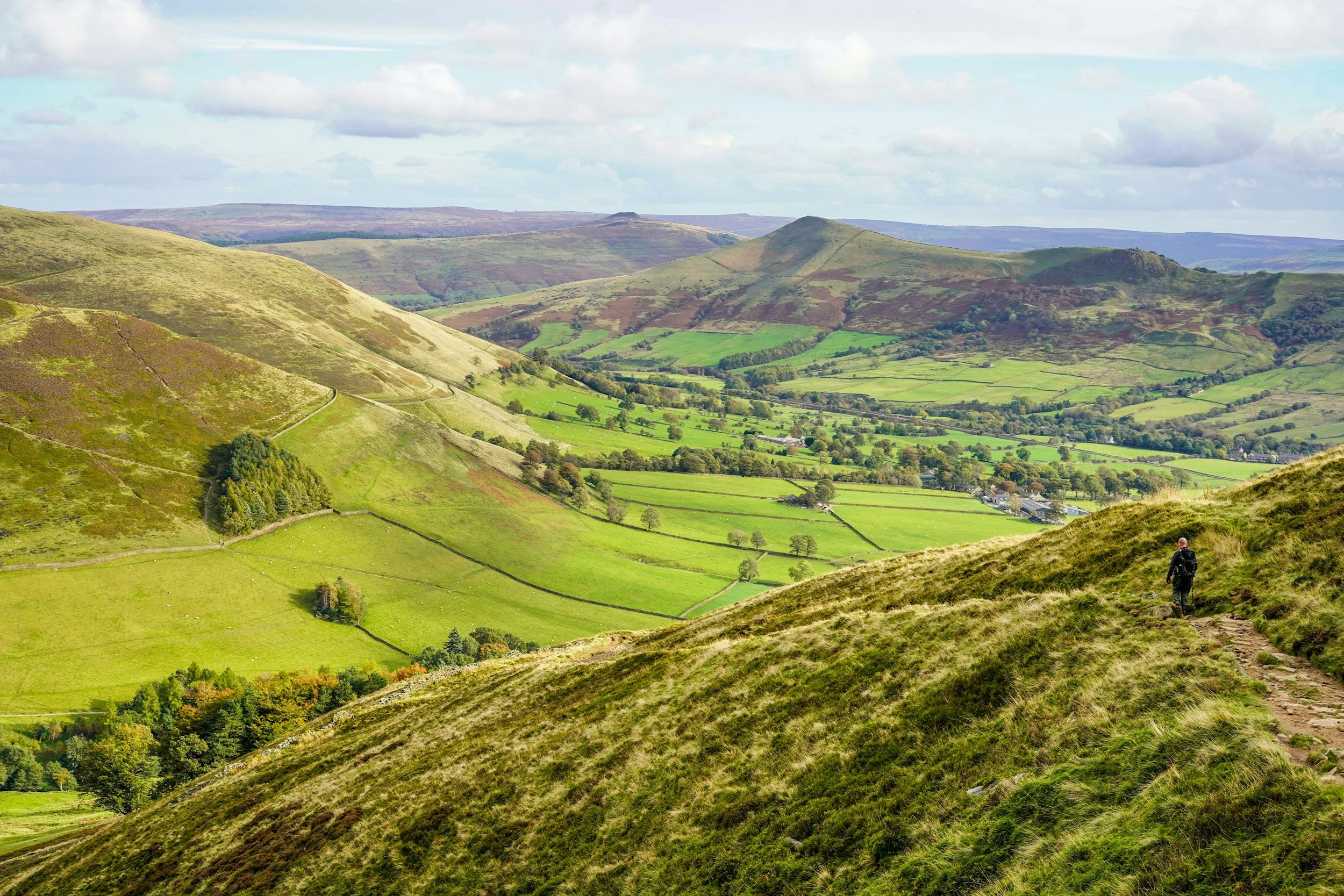Hiker walking on a trail in rolling green hills with farmland and scattered trees, under a partly cloudy sky.