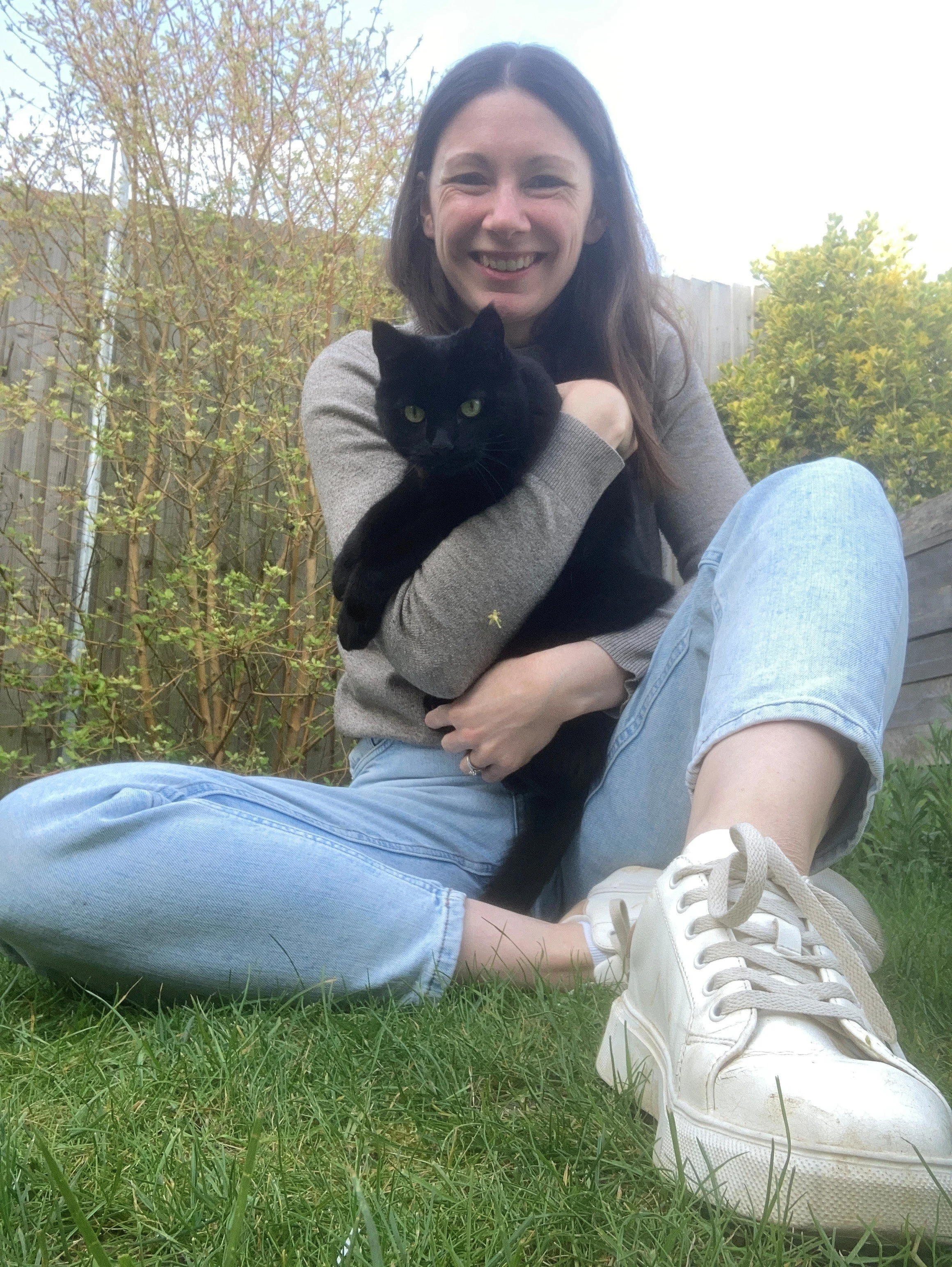A woman with long brown hair smiling while holding a black cat outdoors on a grassy area, with trees and a wooden fence in the background.
