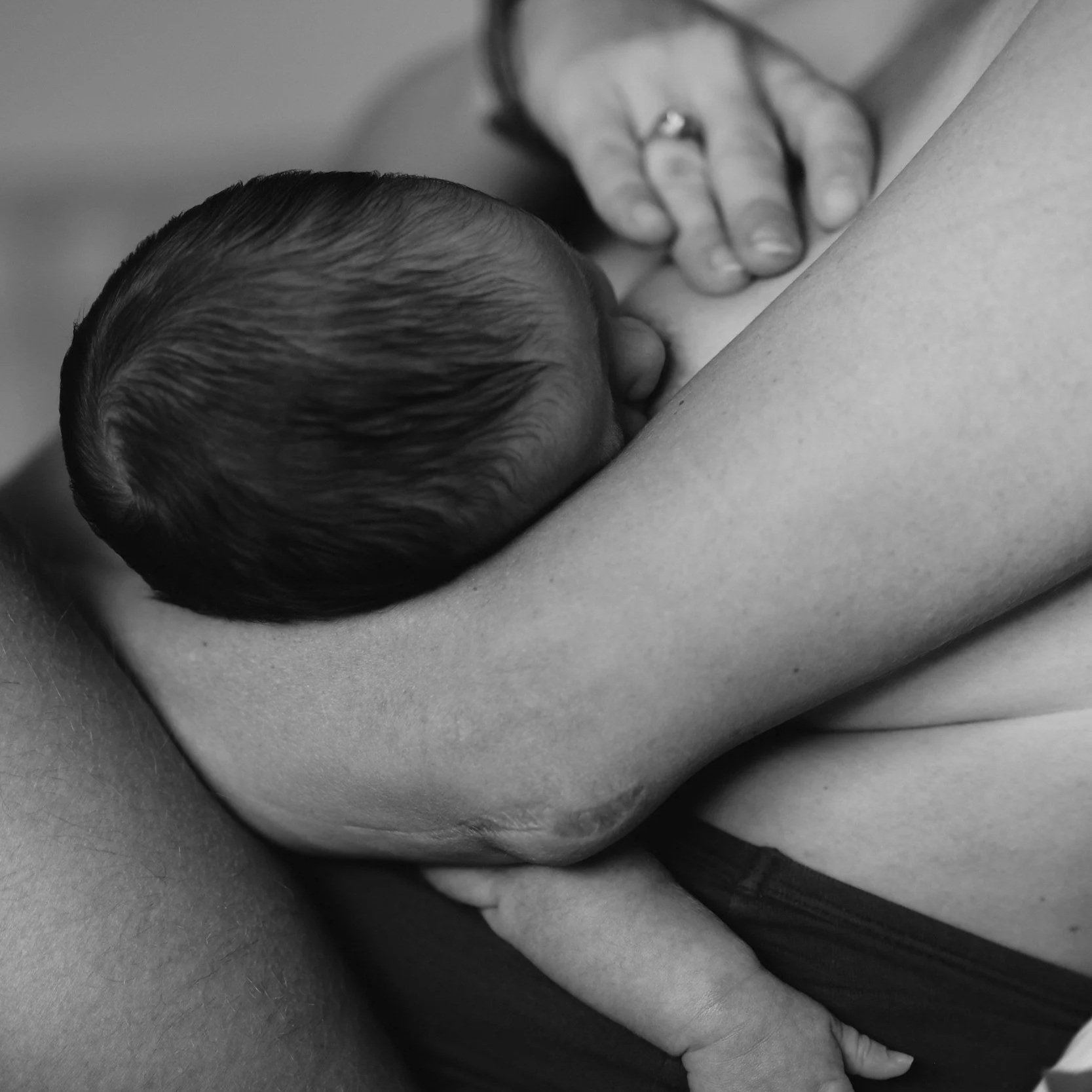 Close-up black and white photo of a newborn baby breastfeeding, with a hand gently placed on the baby's head.