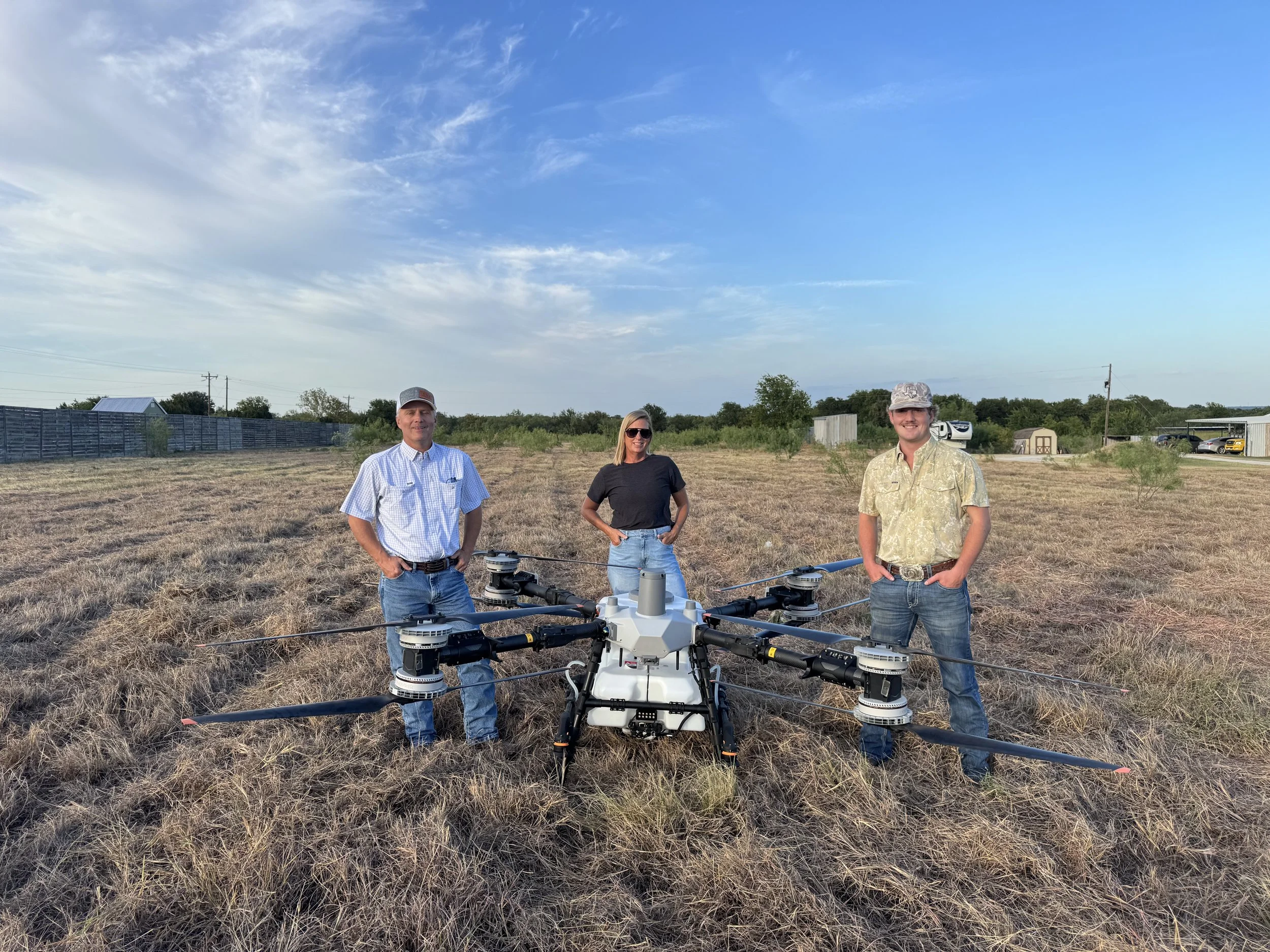 Three people stand outdoors in a field with dry grass, with a drone in front of them. The man on the left wears a light blue shirt and a cap. The woman in the middle wears sunglasses and a dark shirt. The man on the right wears a camouflage hat and a patterned shirt. The sky is mostly clear with some clouds, and there are trees, a fence, and buildings in the background.