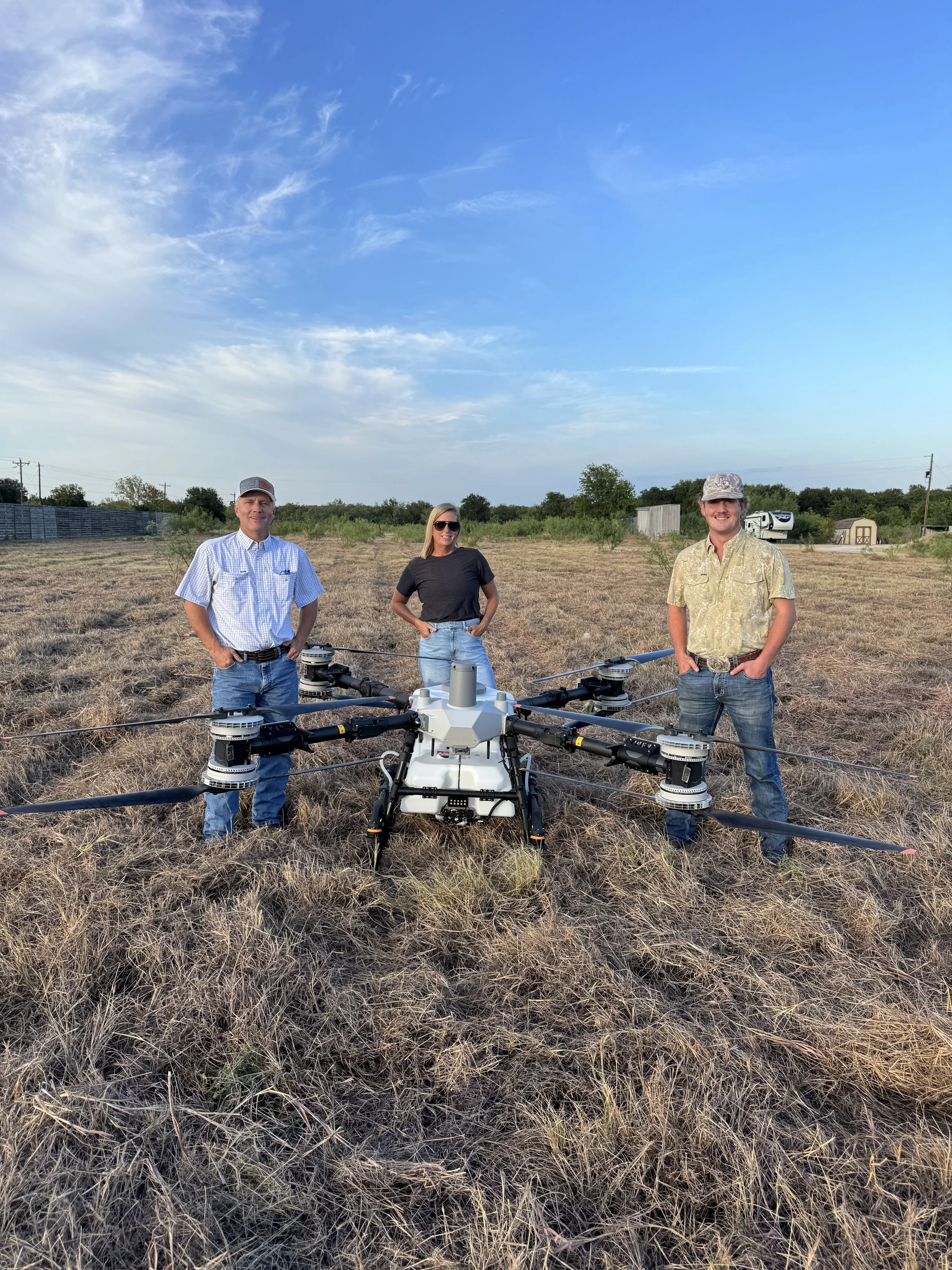 Three people standing outdoors in a field with a drone in the foreground. The man on the left is wearing a checkered shirt and a cap, the woman in the middle is wearing sunglasses and a dark t-shirt, and the man on the right is wearing a patterned shirt and a cap. The background features a clear sky, some trees, and structures like sheds and a camper.