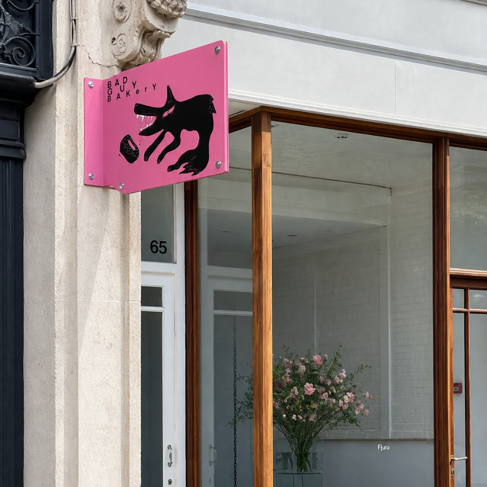 Pink sign with black graphic of a dog and bakery name 'Bad Dog Bakery' outside storefront with large window showing pink flowers inside.