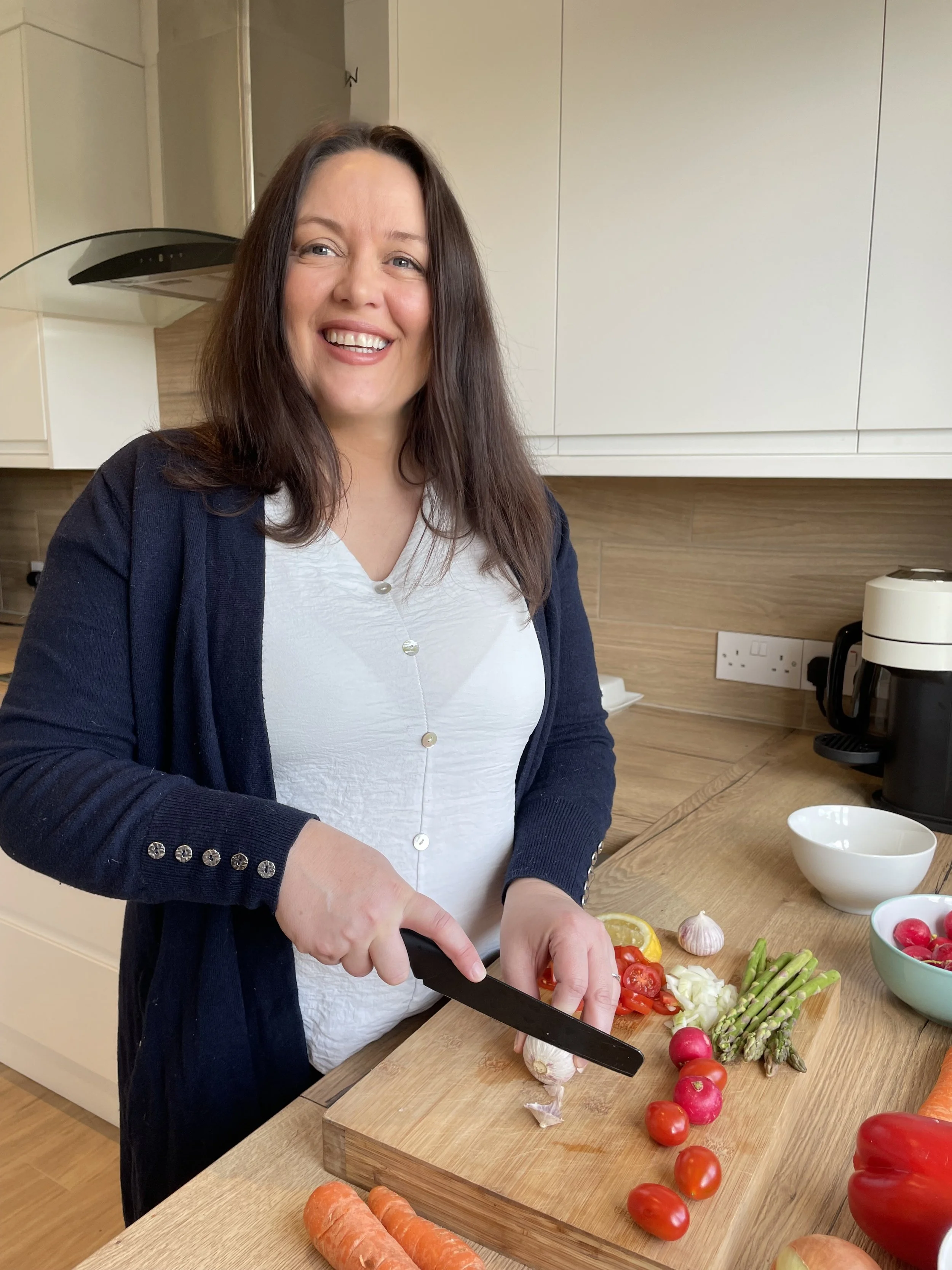 Claire is wearing a white button-up shirt and a navy cardigan, is chopping vegetables on a wooden cutting board in a modern kitchen. The counter has cherry tomatoes, garlic, green asparagus, and other vegetables.