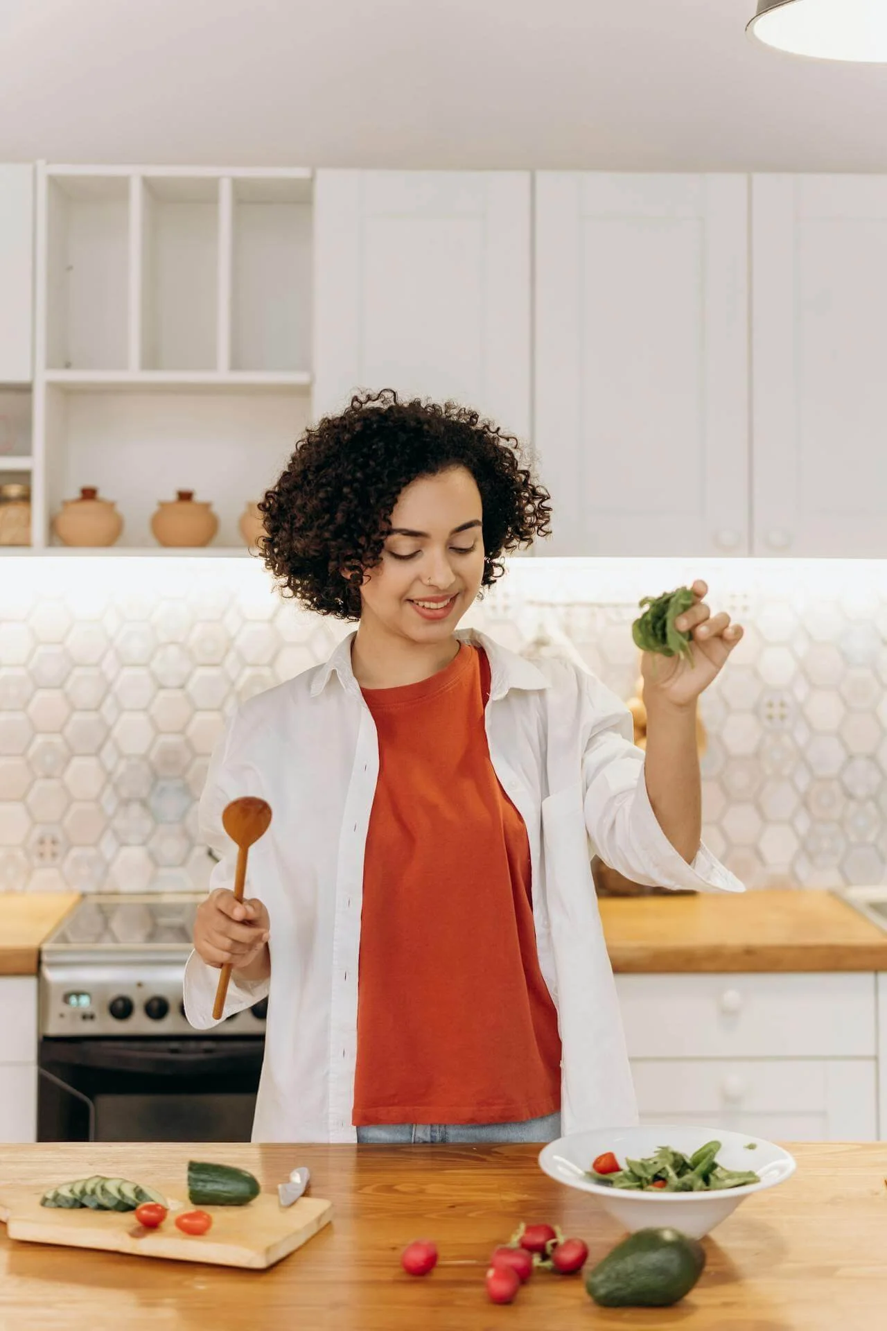 A woman with curly hair, wearing a white shirt over an orange T-shirt, stands in a kitchen preparing a salad. She is holding a bunch of greens in one hand and a wooden spoon in the other. On the wooden kitchen counter, there are cut vegetables such as cucumbers, radishes, and cherry tomatoes, along with a bowl of salad and a cutting board.