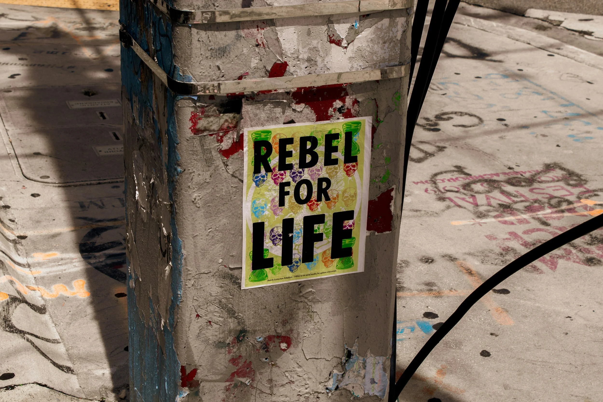 Close-up of a weathered, concrete utility pole with peeling paint and a colorful poster that reads 'Rebel for Life' with skull illustrations, situated on a graffiti-covered sidewalk.
