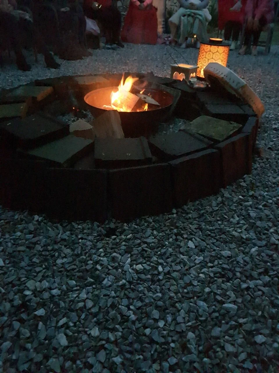 Outdoor gathering around a fire pit with people sitting in the background during twilight or evening.