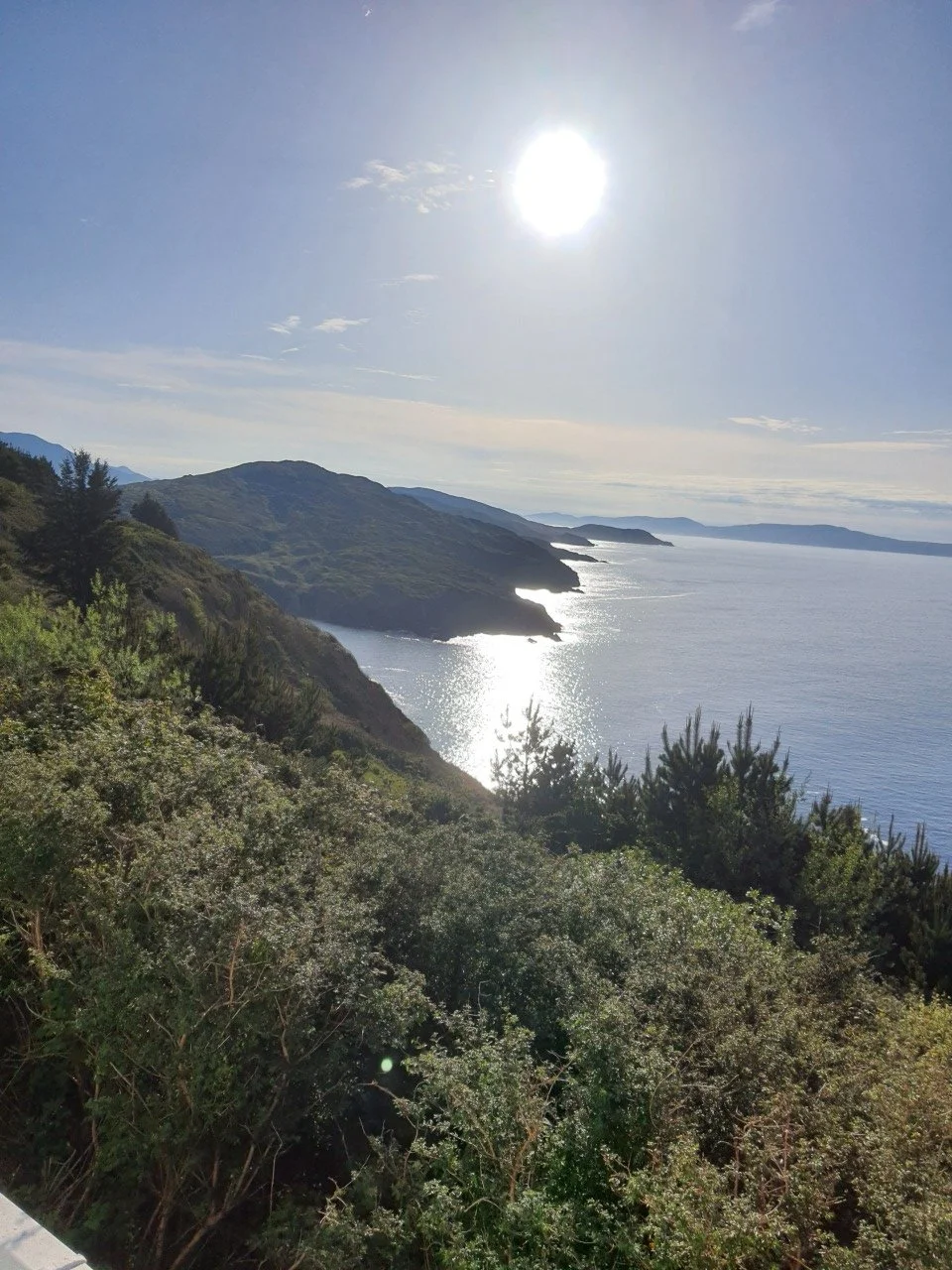 Scenic view of a coastal landscape showing the ocean, green hills, and trees, with the sun shining brightly in the sky.