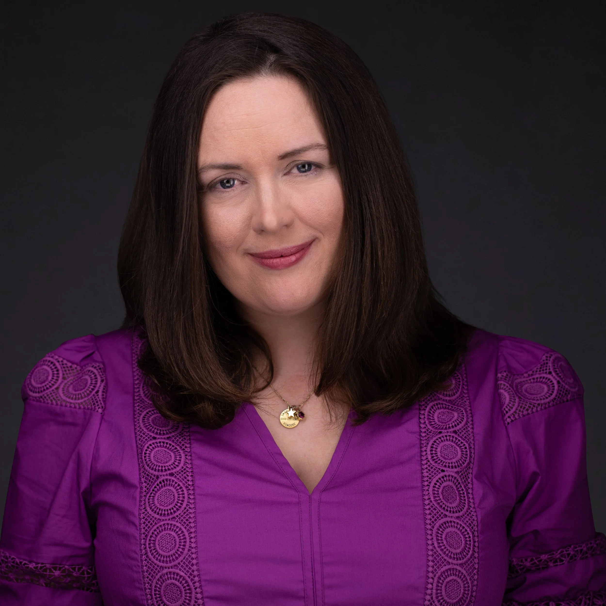 Claire OBrien , wearing a purple top with intricate circular embroidery, a gold necklace with a star-shaped pendant, and smiling against a dark background.