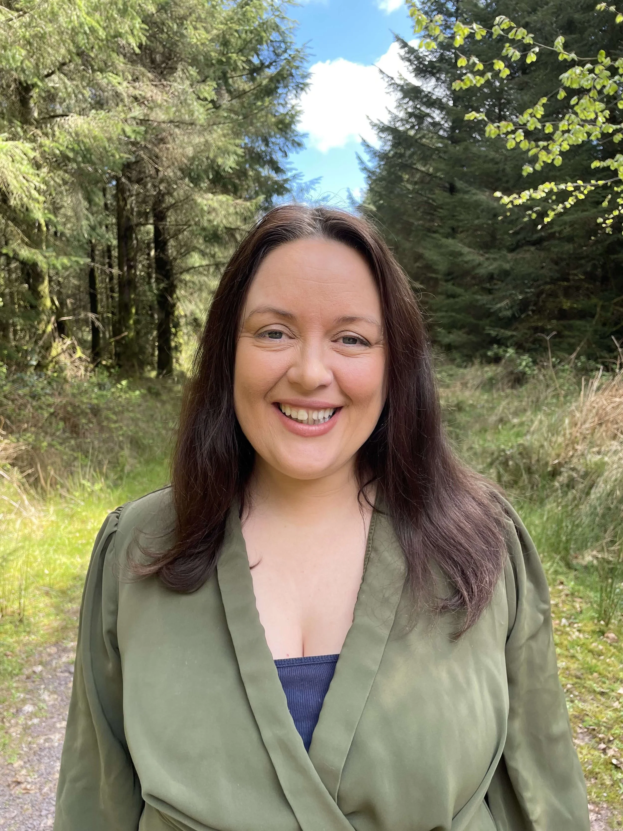 A woman with long brown hair, smiling, wearing an olive-green jacket over a blue top, standing outdoors in a forested area with green trees and a blue sky in the background.