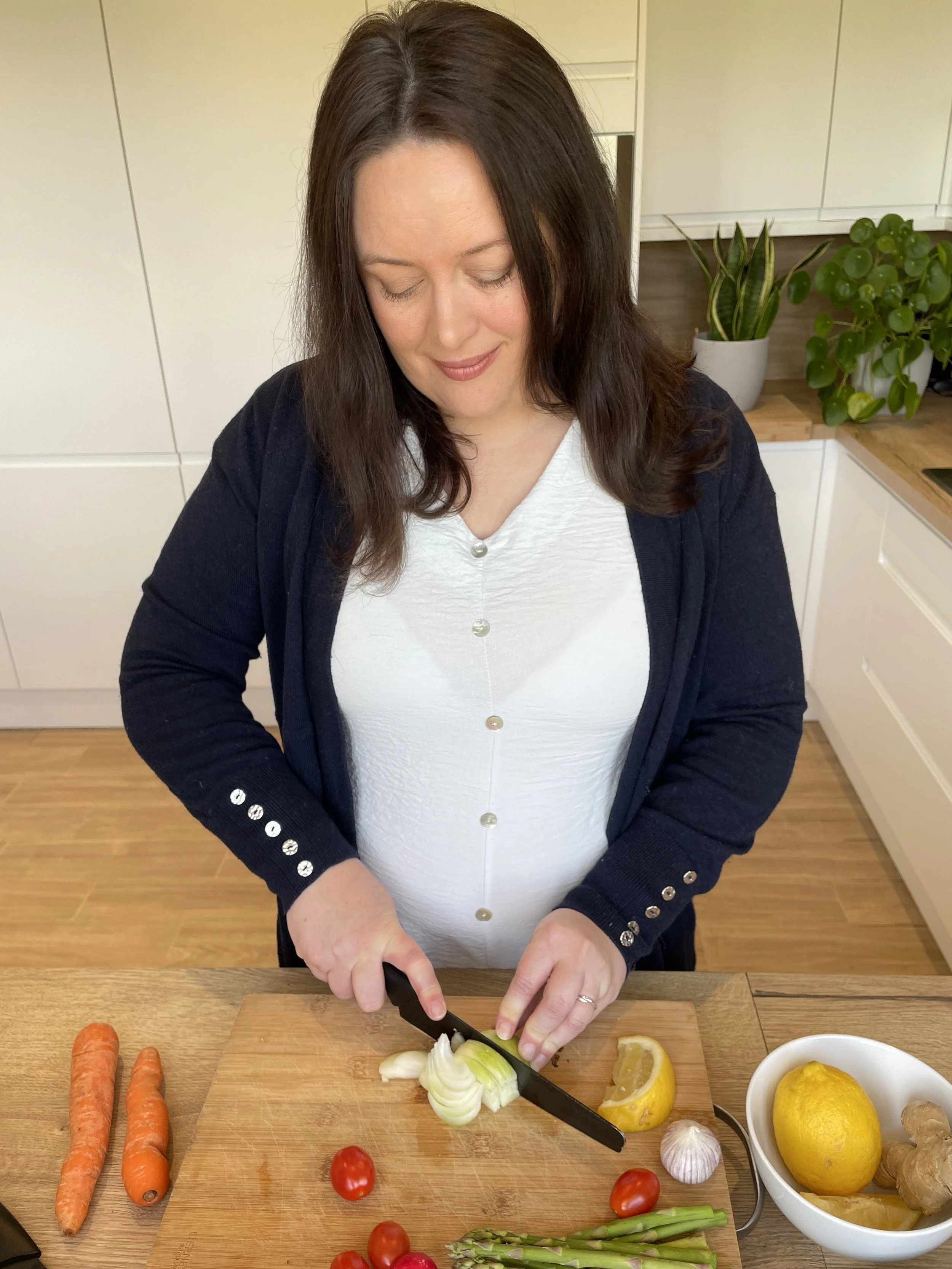A woman chopping onions on a wooden cutting board with vegetables and lemons nearby in a kitchen.